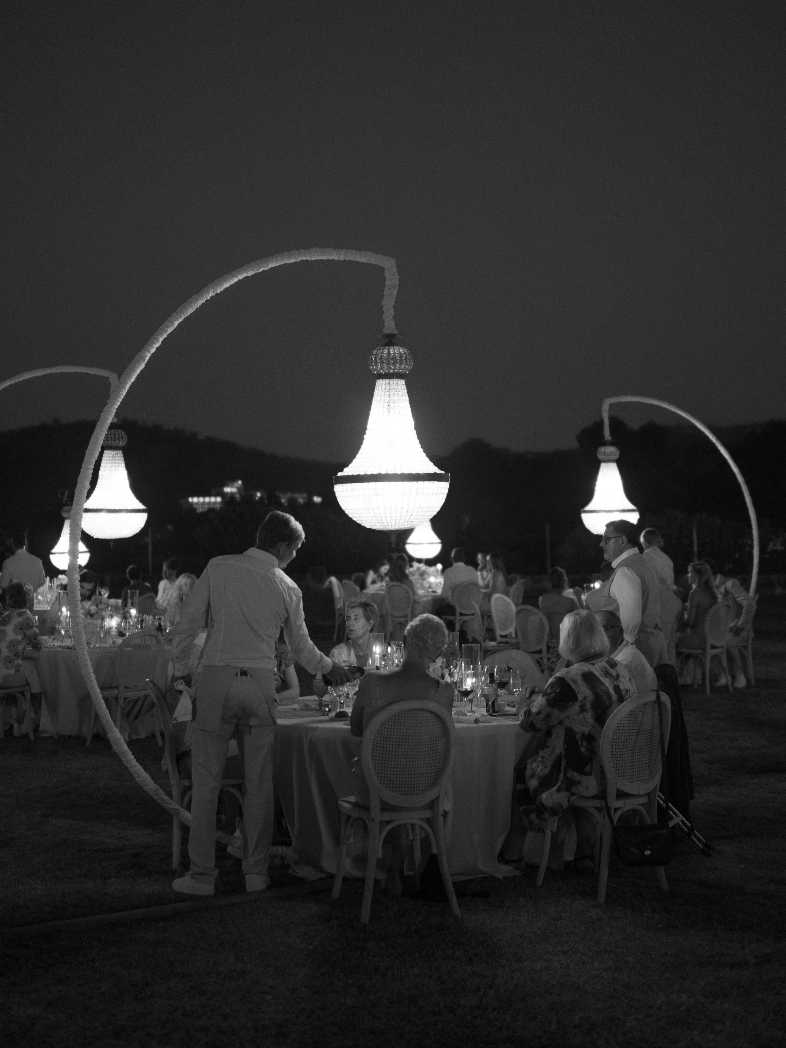 A black and white photo of an outdoor dinner event at night, featuring large illuminated chandeliers hanging above round tables with seated guests, and waitstaff serving food.