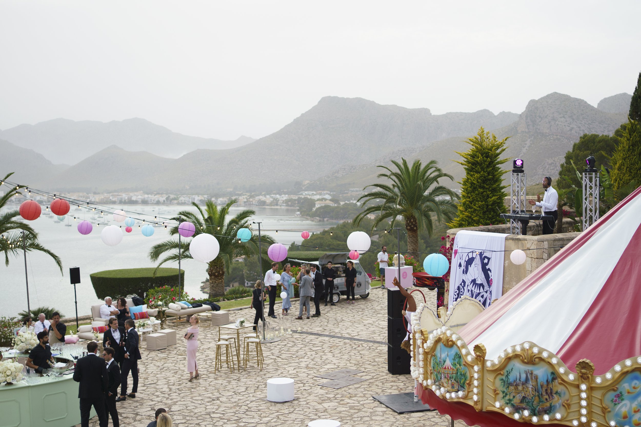 Outdoor celebration with palm trees, colorful lanterns, and a mountain landscape in the background. People are gathering and a DJ is playing music on a stage. There is a carousel ride in the foreground.