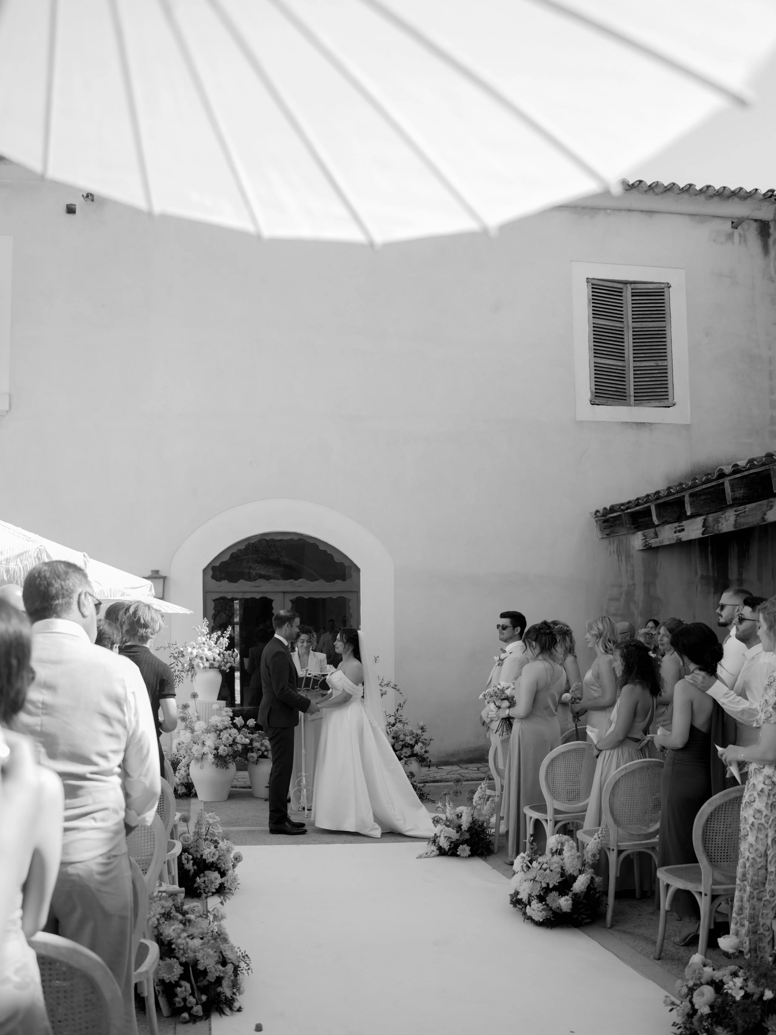 A wedding ceremony outdoors with a bride and groom exchanging vows in front of their guests, with floral arrangements and decorated chairs.