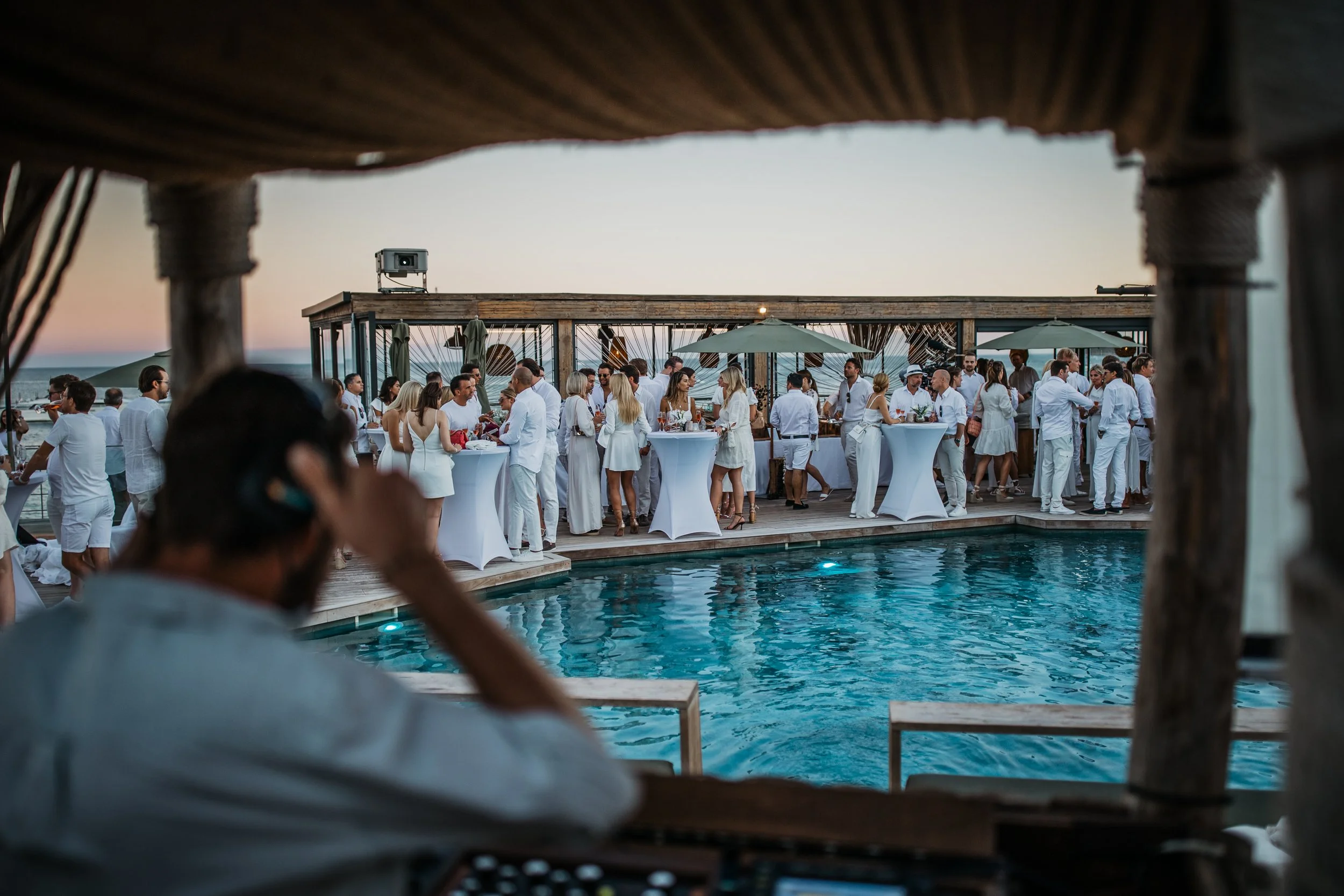 Guests in white attire enjoying a social event by a pool at sunset, on a rooftop with umbrellas and wooden decor.