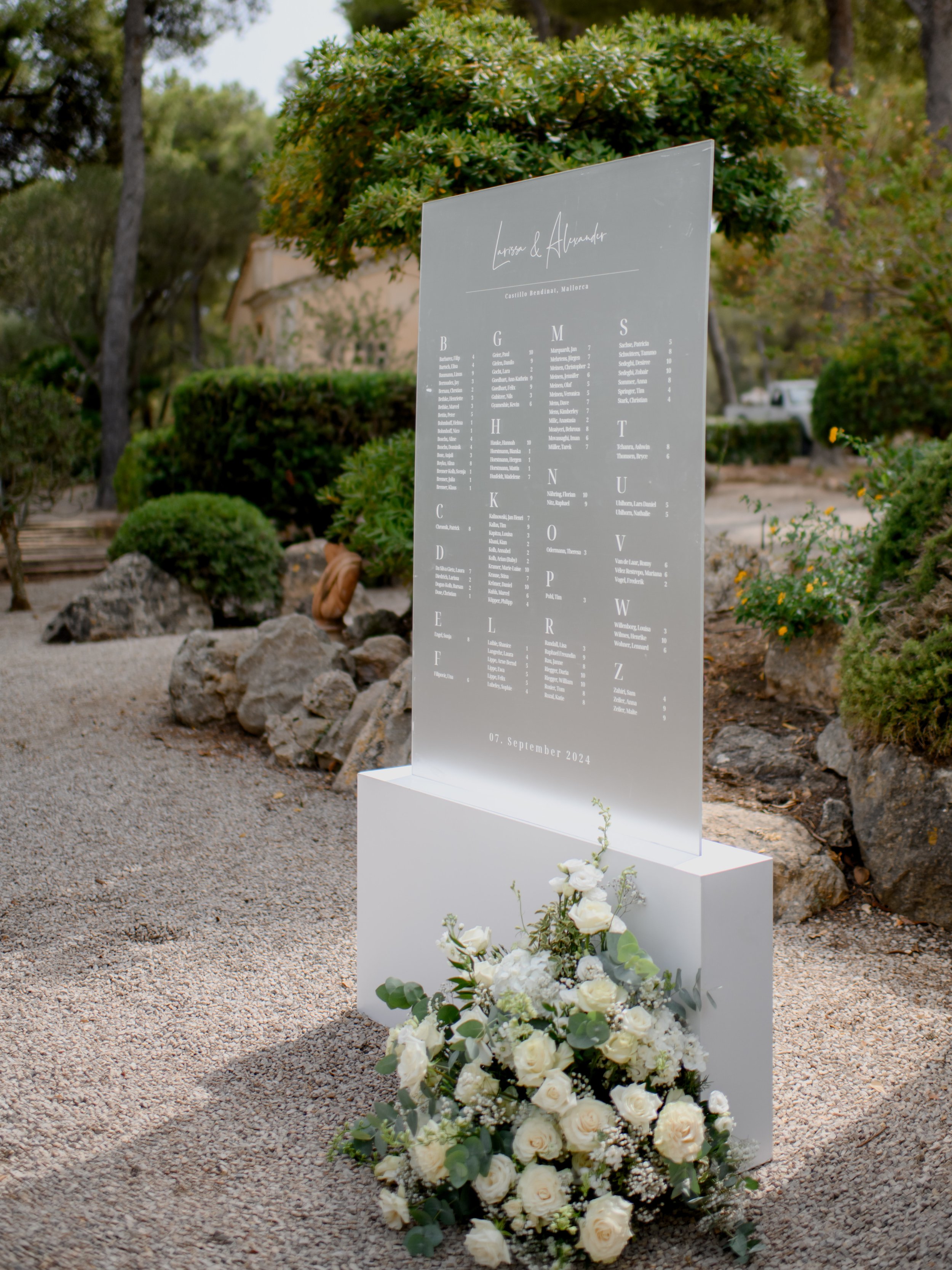 A wedding seating chart displayed on a clear acrylic board with white text, decorated with a bouquet of white roses at the base, set outdoors with trees and bushes in the background.