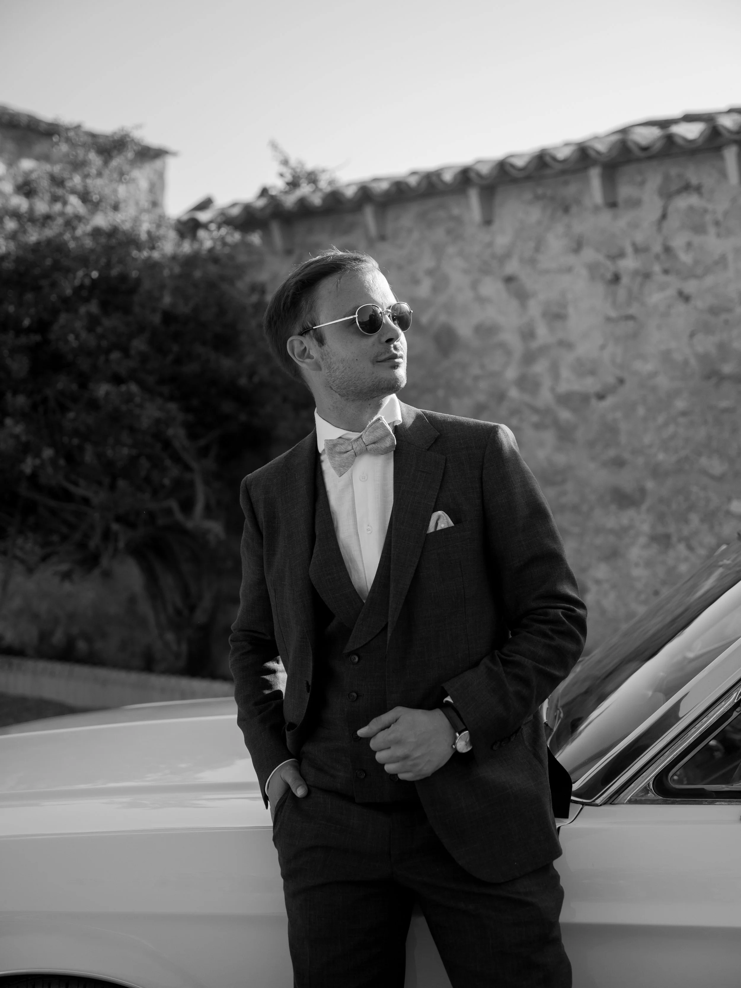 A man in a suit and sunglasses leans against a vintage car outdoors with a stone wall and trees in the background.