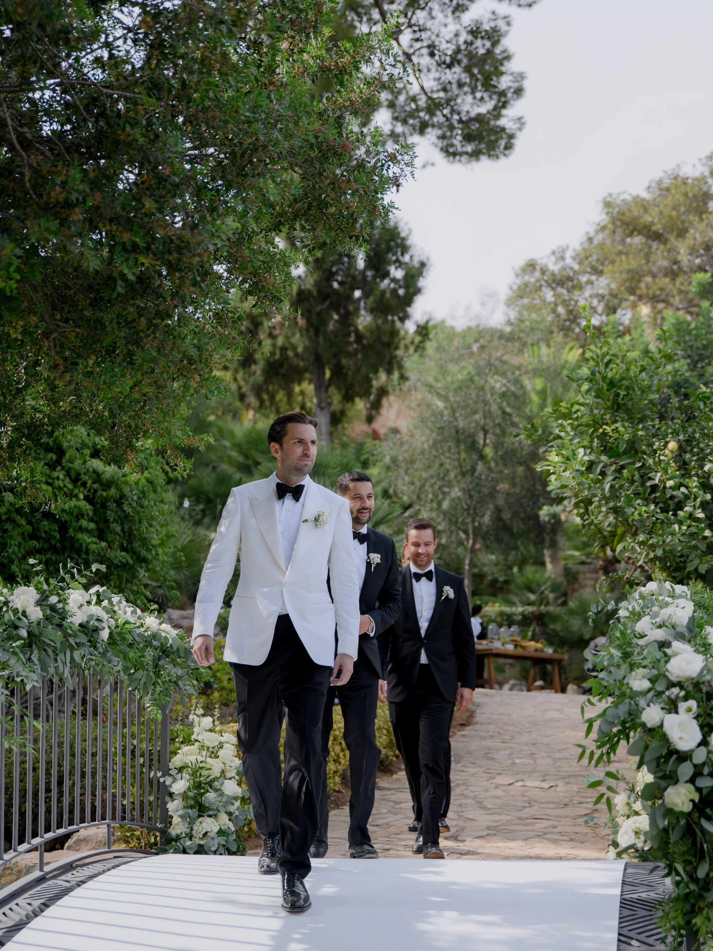 A group of three men in tuxedos walking on a white aisle at an outdoor wedding ceremony, surrounded by greenery and white floral arrangements.
