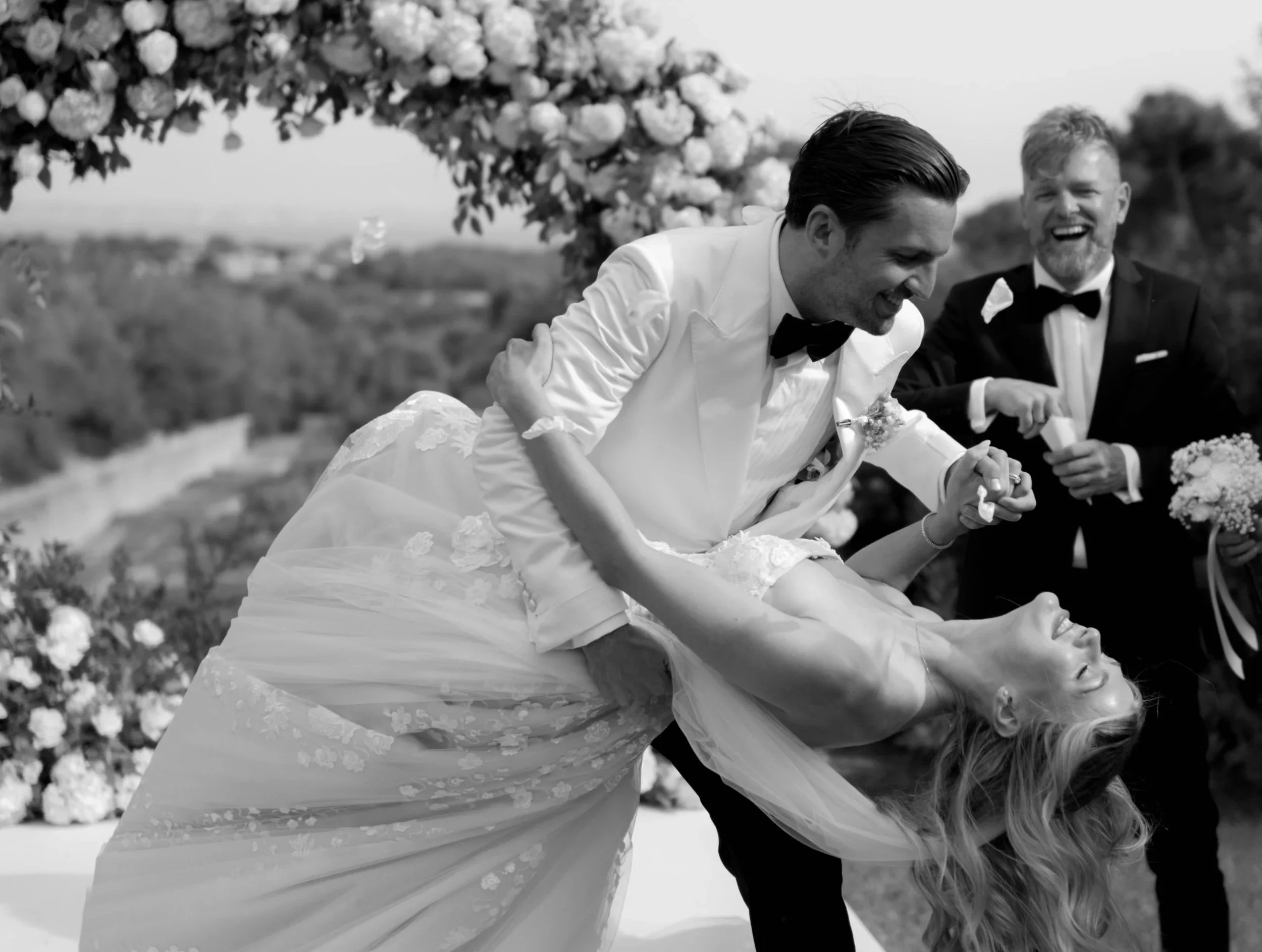 A black and white photo of a wedding scene with a groom dipping and kissing the bride, who is looking up with her eyes closed. The groom is wearing a white tuxedo with a black bow tie, and the bride is in a lace wedding dress. Another man in a black tuxedo is standing behind them, smiling and holding a bouquet of flowers. The background features a scenic outdoor setting with trees and cloudy sky.