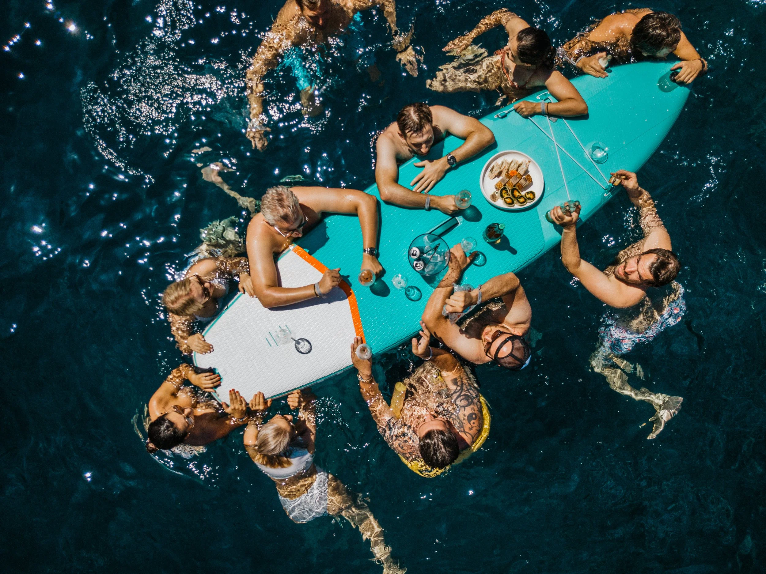 Group of people floating and sitting on a large turquoise paddleboard in the water, enjoying drinks and snacks during a sunny day.