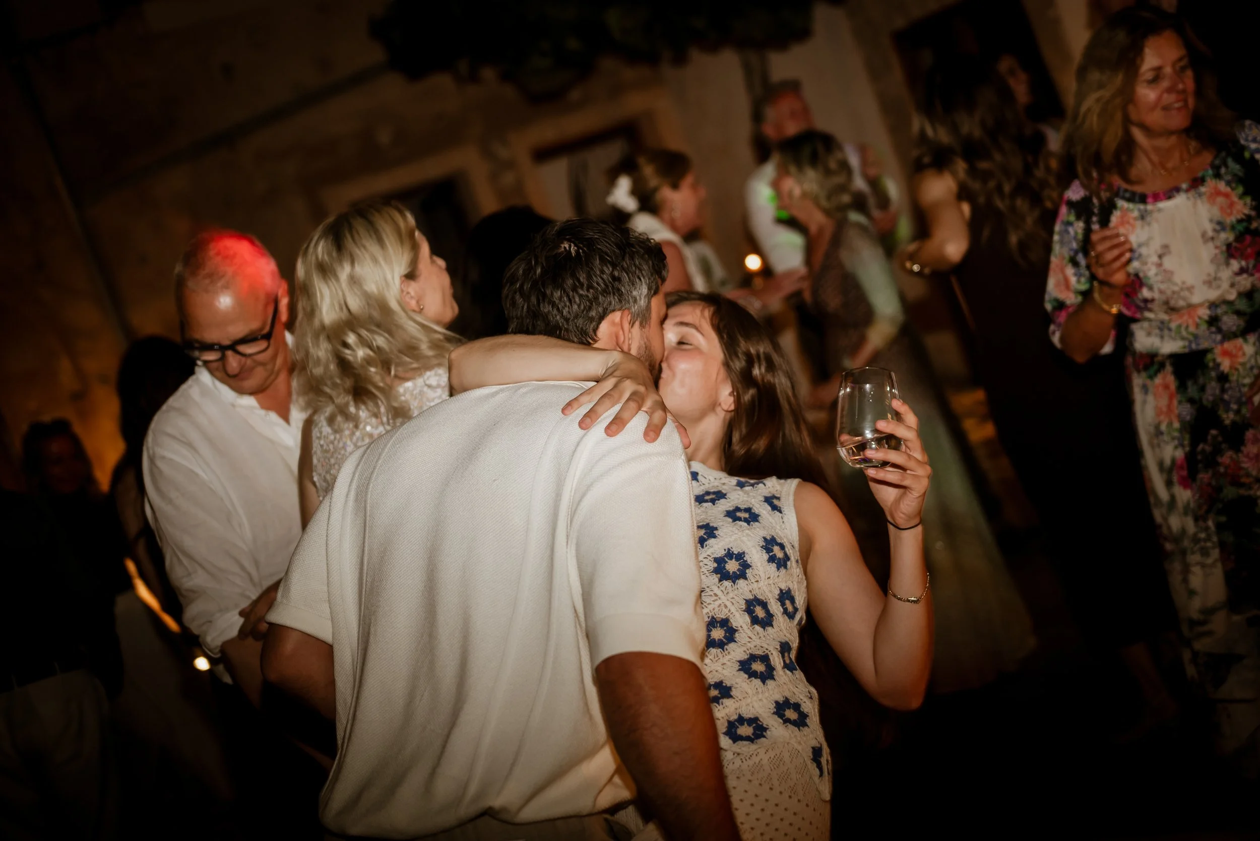 Couple kissing at a lively indoor party with other guests dancing and socializing in the background.