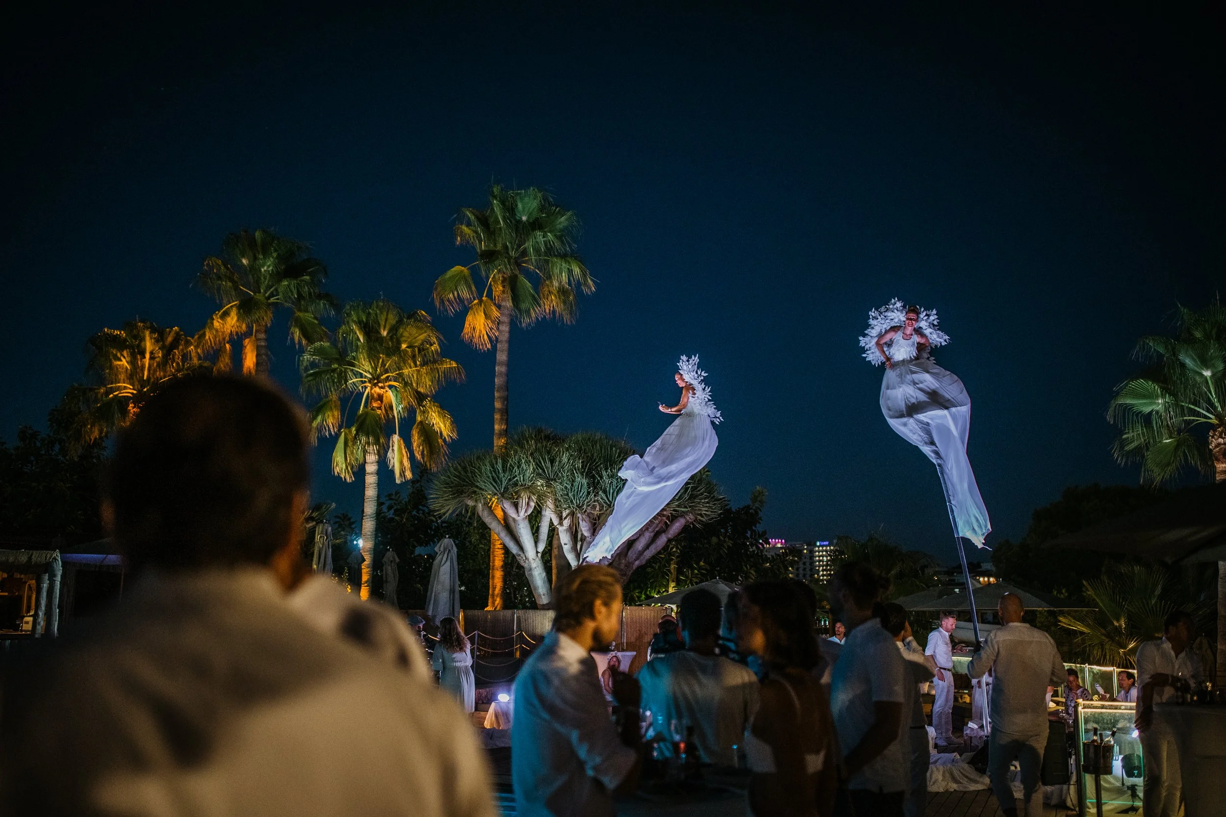 Nighttime outdoor event with illuminated dancers on stilts in flowing white costumes and floral headpieces, surrounded by palm trees and spectators at a tropical venue.