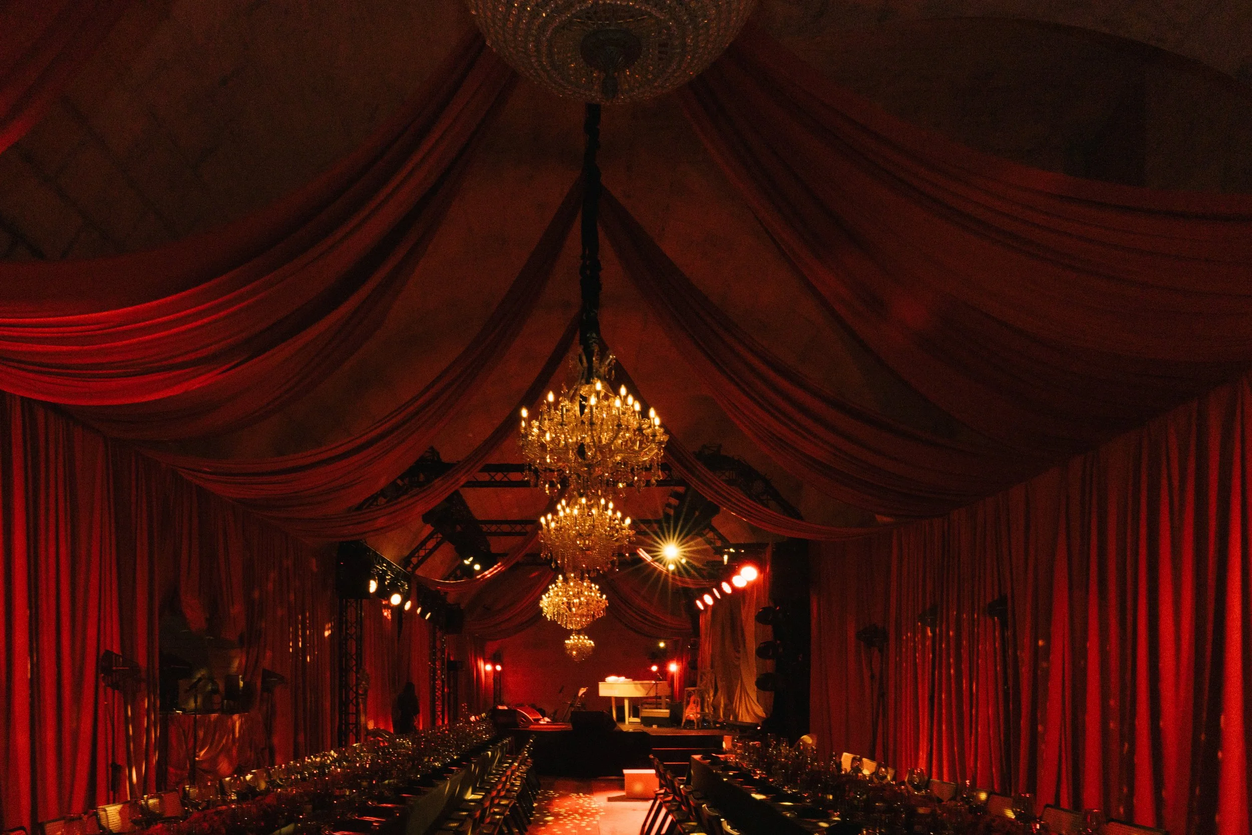 Elegant event hall decorated with red drapery, chandeliers, and lighting, with tables set for a formal dinner and a stage at the end.