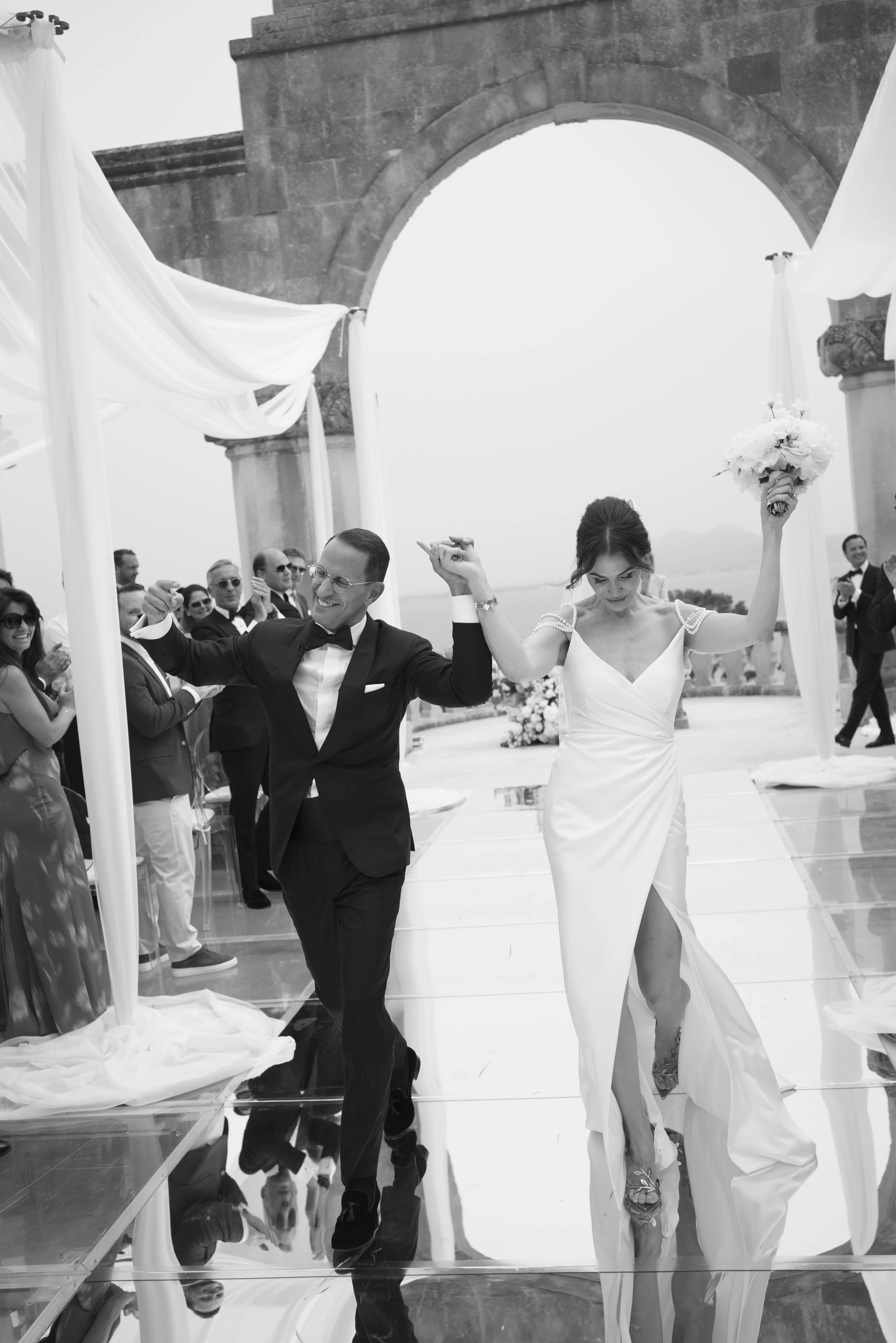 A newly married couple celebrating on their wedding day, walking hand-in-hand under a stone archway with guests clapping in the background.