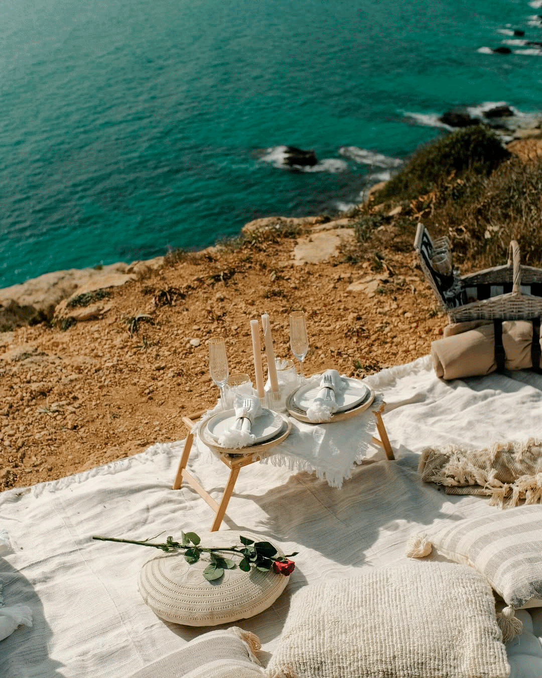 A romantic picnic setup on a blanket overlooking the ocean with a small table set with two plates, glasses, and candles, surrounded by cushions and a chair, on a rocky cliffside.
