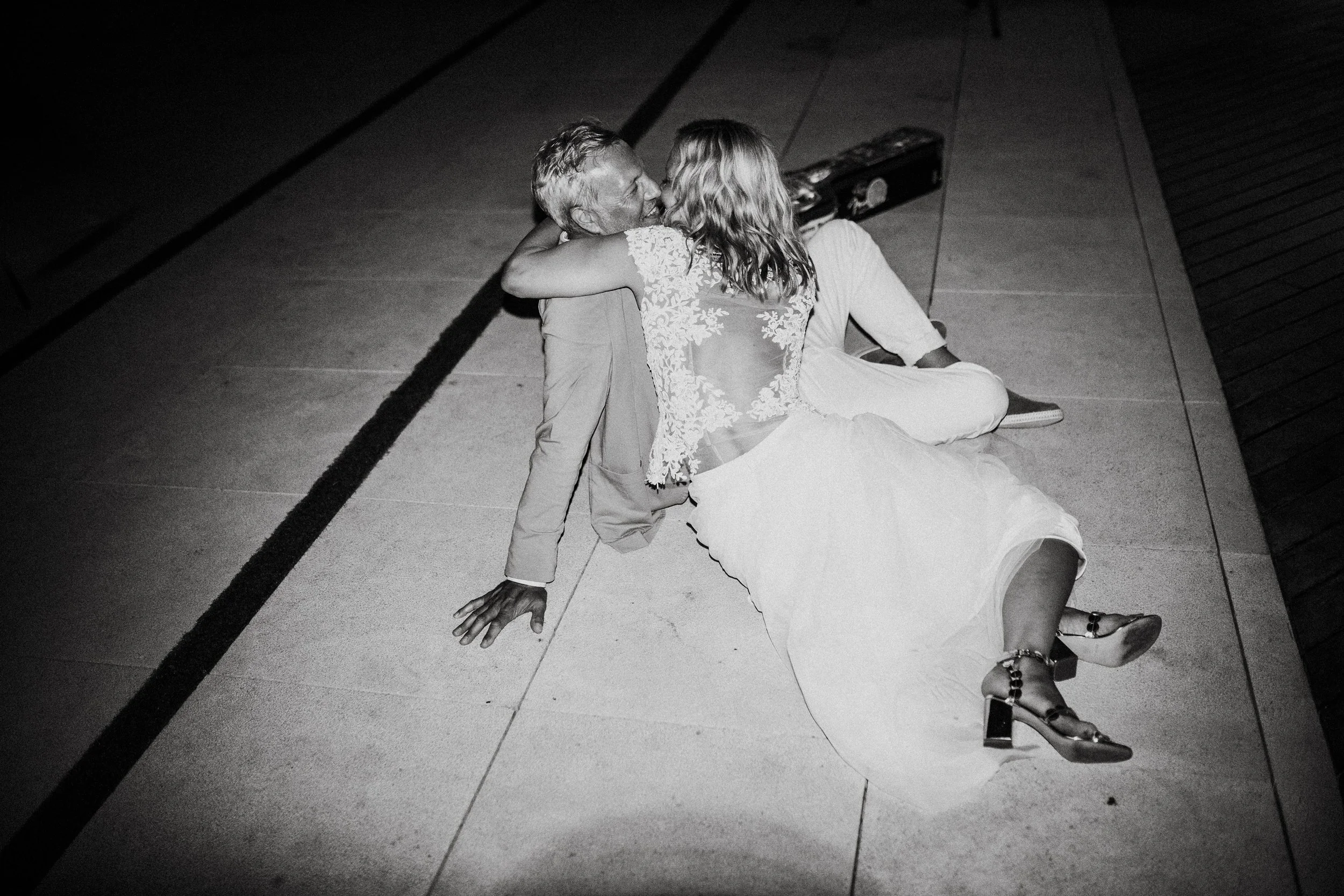 A black and white photo of a man and woman sharing a kiss on the floor. The woman is wearing a lace dress and high heels, while the man is in a suit. They are sitting on tile flooring with a violin case nearby.