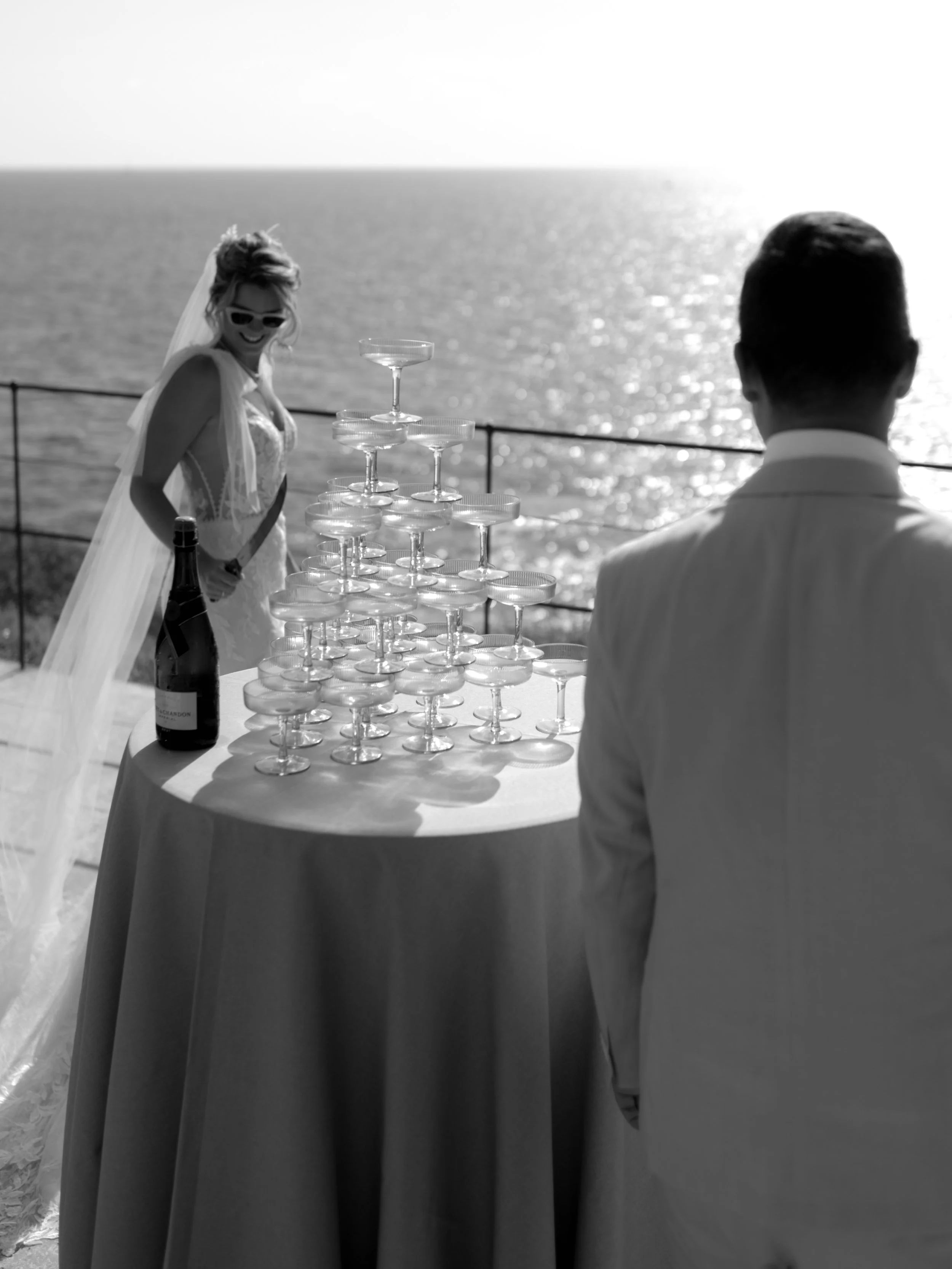 A bride and groom at an outdoor wedding by the water, with a tower of champagne glasses on a table between them.