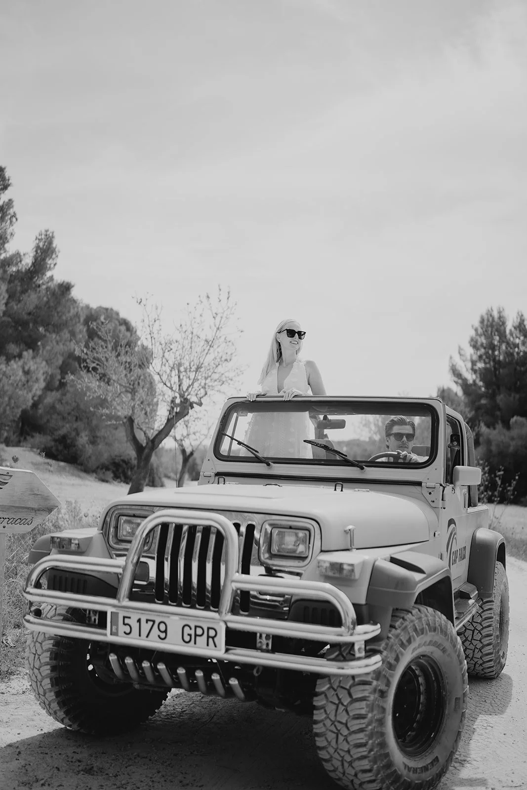 A black-and-white photo of a woman standing in an open-top off-road vehicle with a man driving, outdoors with trees and a partly cloudy sky in the background.