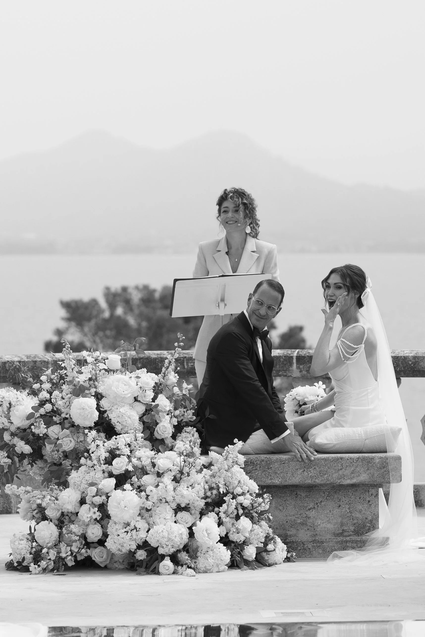 A wedding ceremony with a bride and groom sitting by a large floral arrangement outdoors, with a mountain and lake in the background. An officiant stands behind them holding a book.