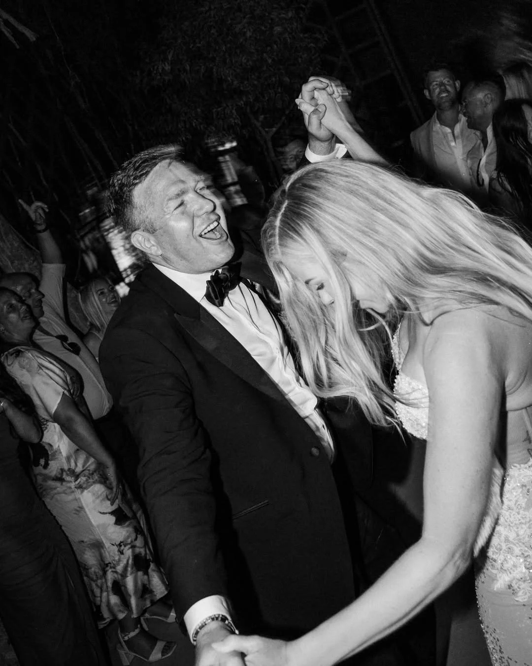 A black and white photo showing a joyful man in a tuxedo and a woman in a wedding dress dancing together at a celebration.
