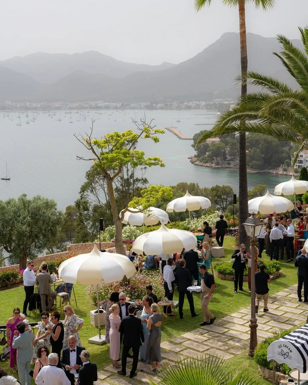 Outdoor social gathering with guests in formal attire, white umbrellas, and lush greenery overlooking a lake with sailboats and mountains in the background.