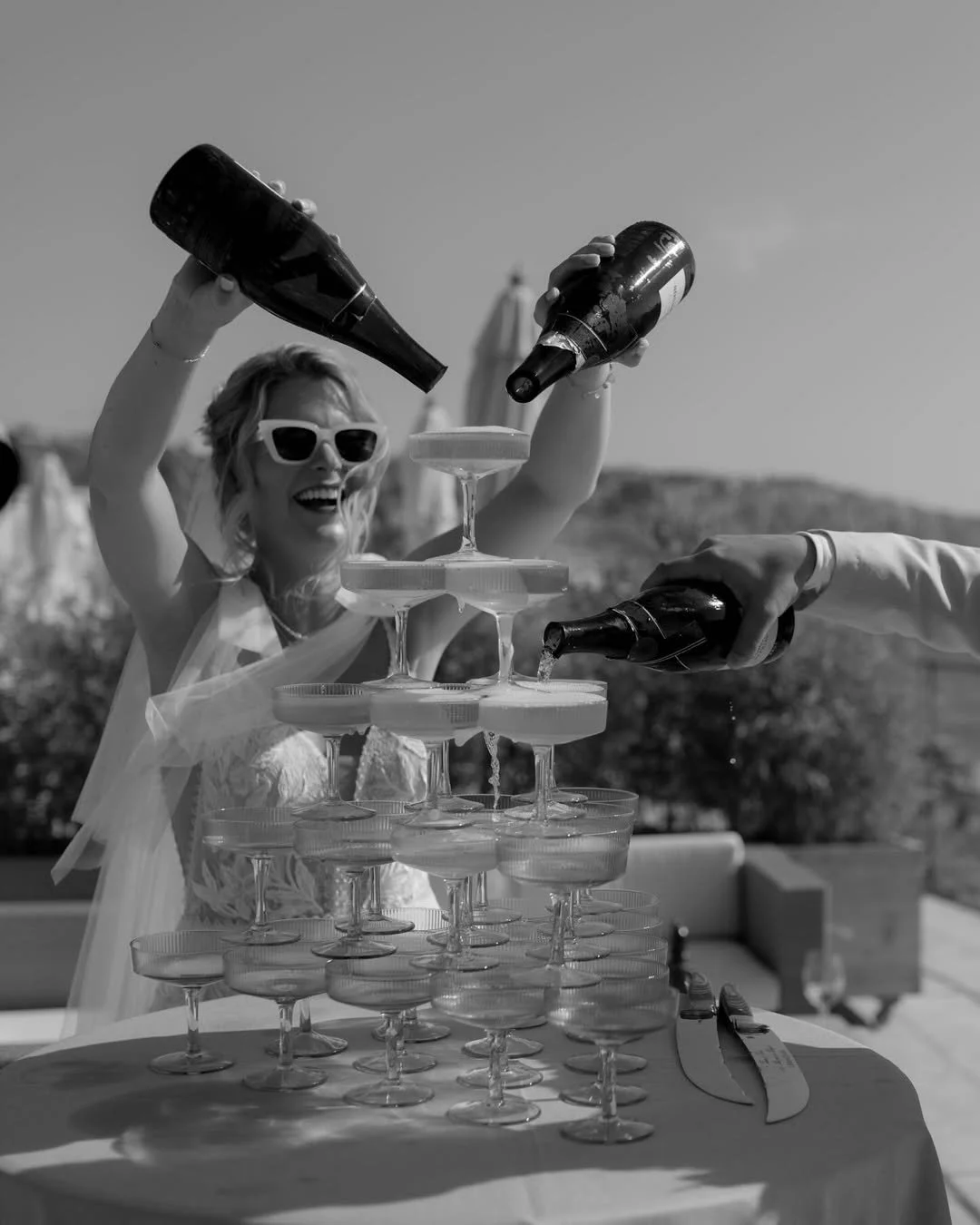 A woman wearing sunglasses celebrating at an outdoor party, pouring champagne into a tower of glasses with another person. She is smiling joyfully amidst an elegant setting.