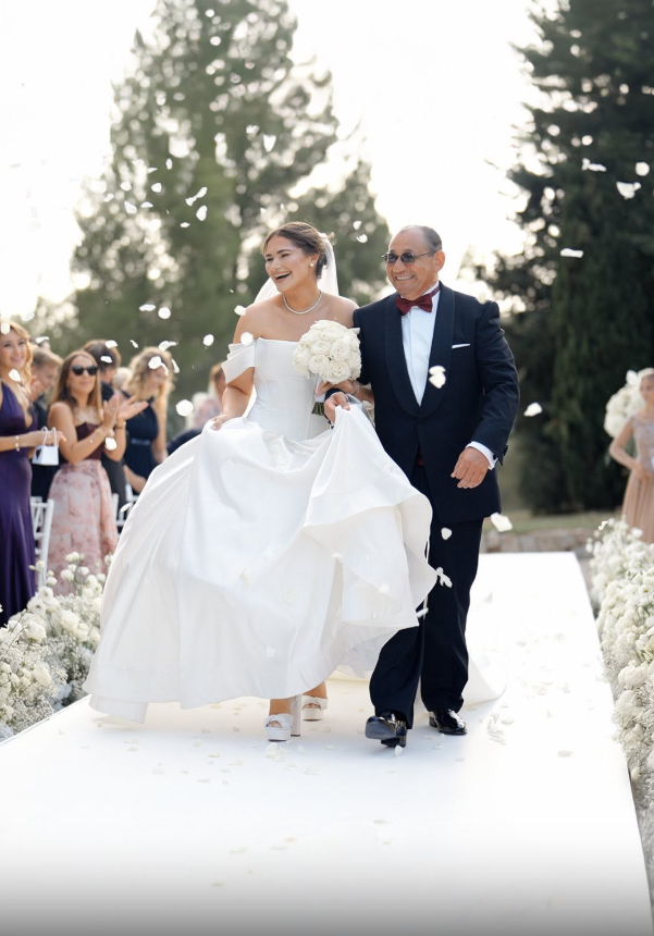 A bride in a white wedding dress holding a bouquet of white roses walking down the aisle with a man in formal attire, surrounded by guests clapping and celebrating outdoors on a sunny day.