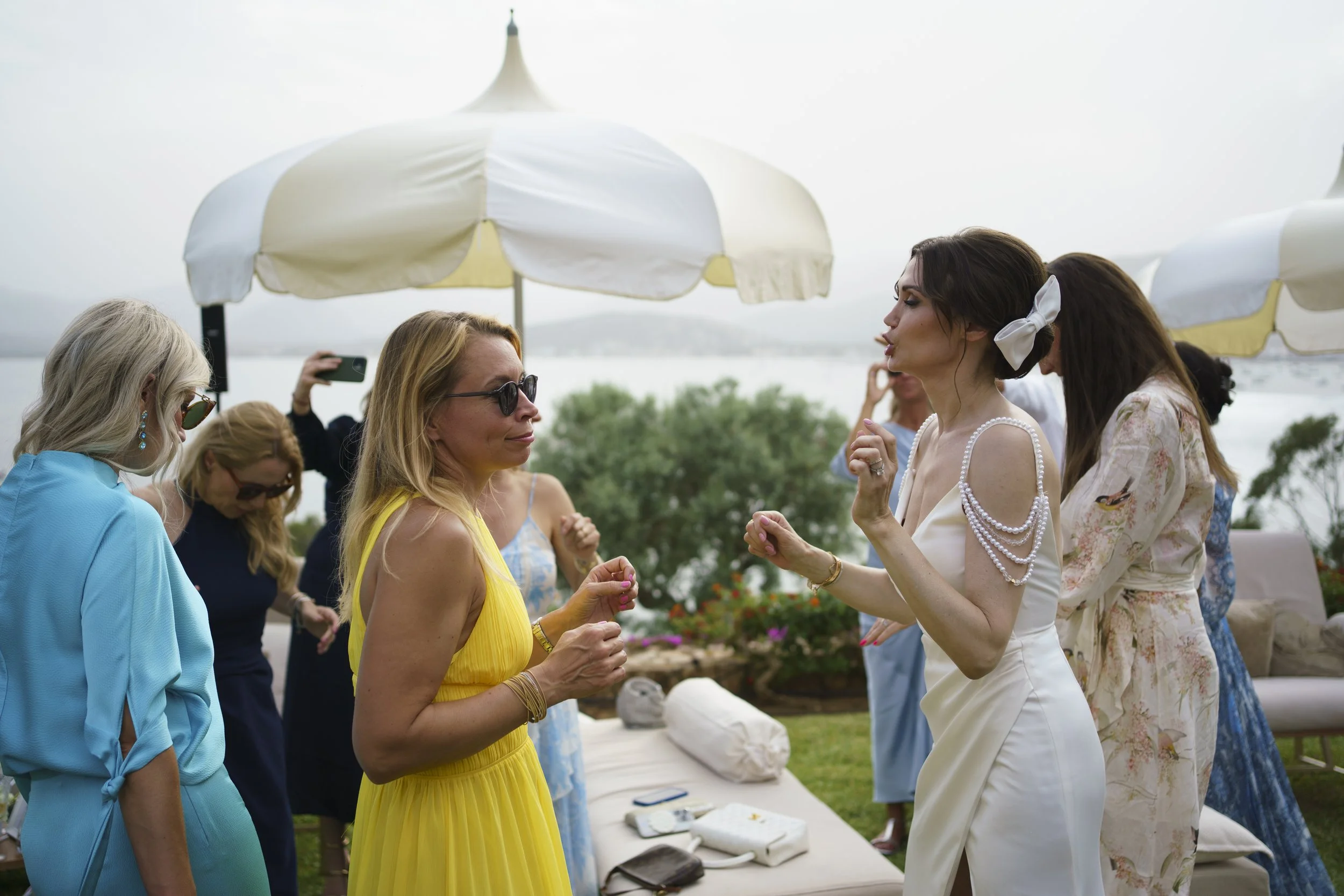 A group of women at an outdoor party or gathering near a body of water, with some wearing colorful dresses and accessories, under umbrellas, engaging in conversation and taking photos.