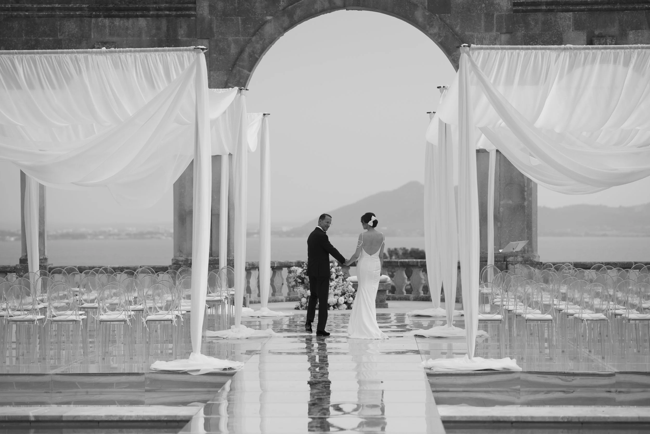 Black and white photo of a wedding ceremony, with a bride and groom holding hands under draped fabric on a terrace overlooking a body of water and mountains.