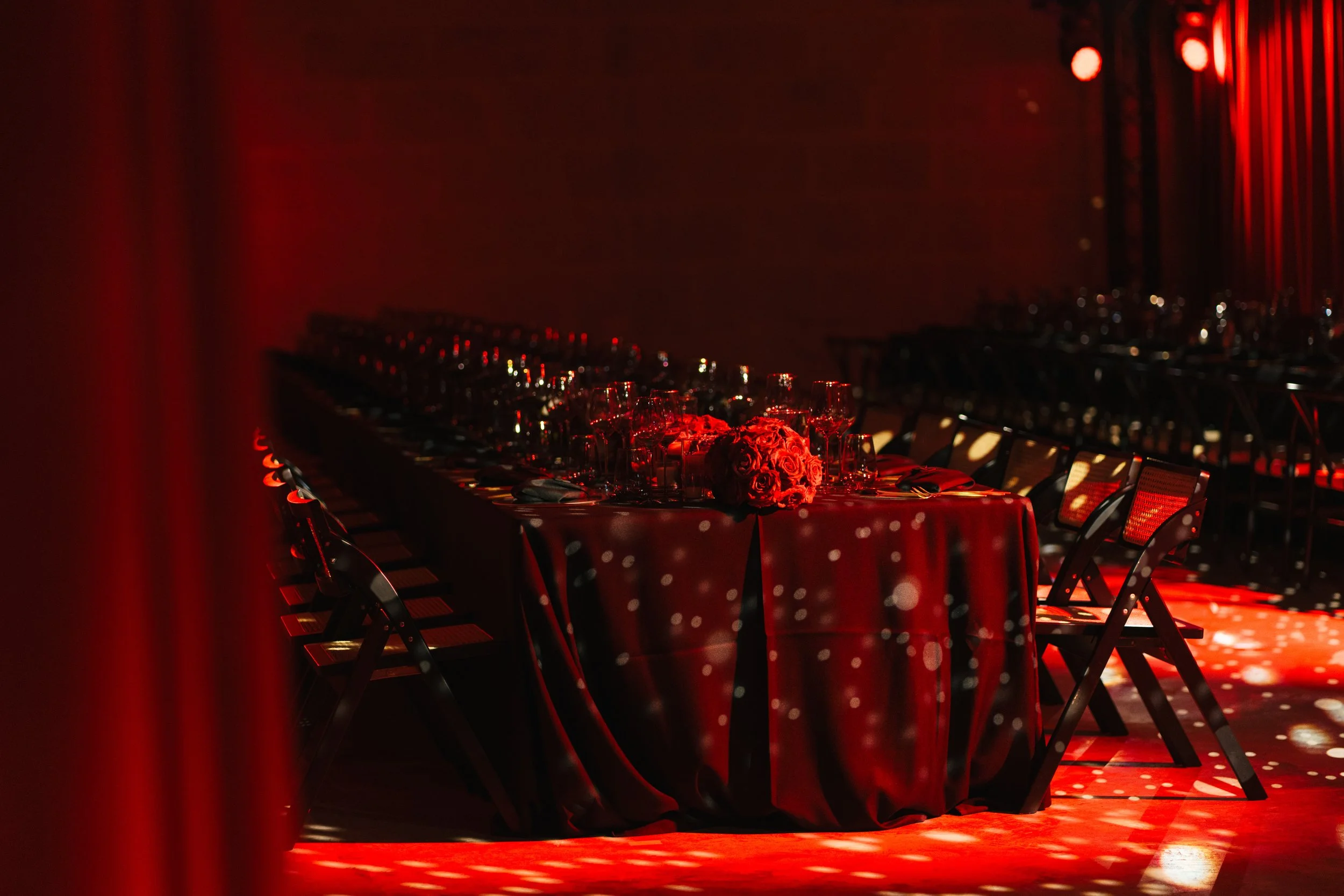 A long banquet table set for a formal event with black chairs, red tablecloth, and a floral centerpiece, illuminated with red lighting