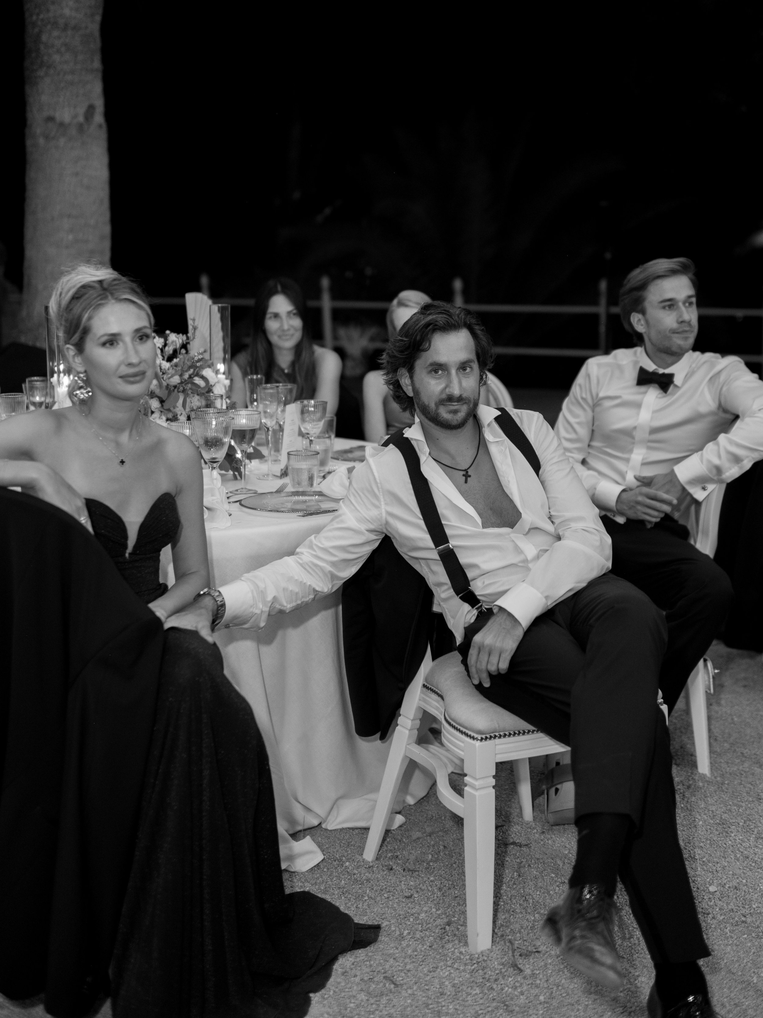 A black and white photo of people seated at a dinner party outdoors at night, with a woman in a strapless dress and a man in a white shirt with suspenders in the foreground.