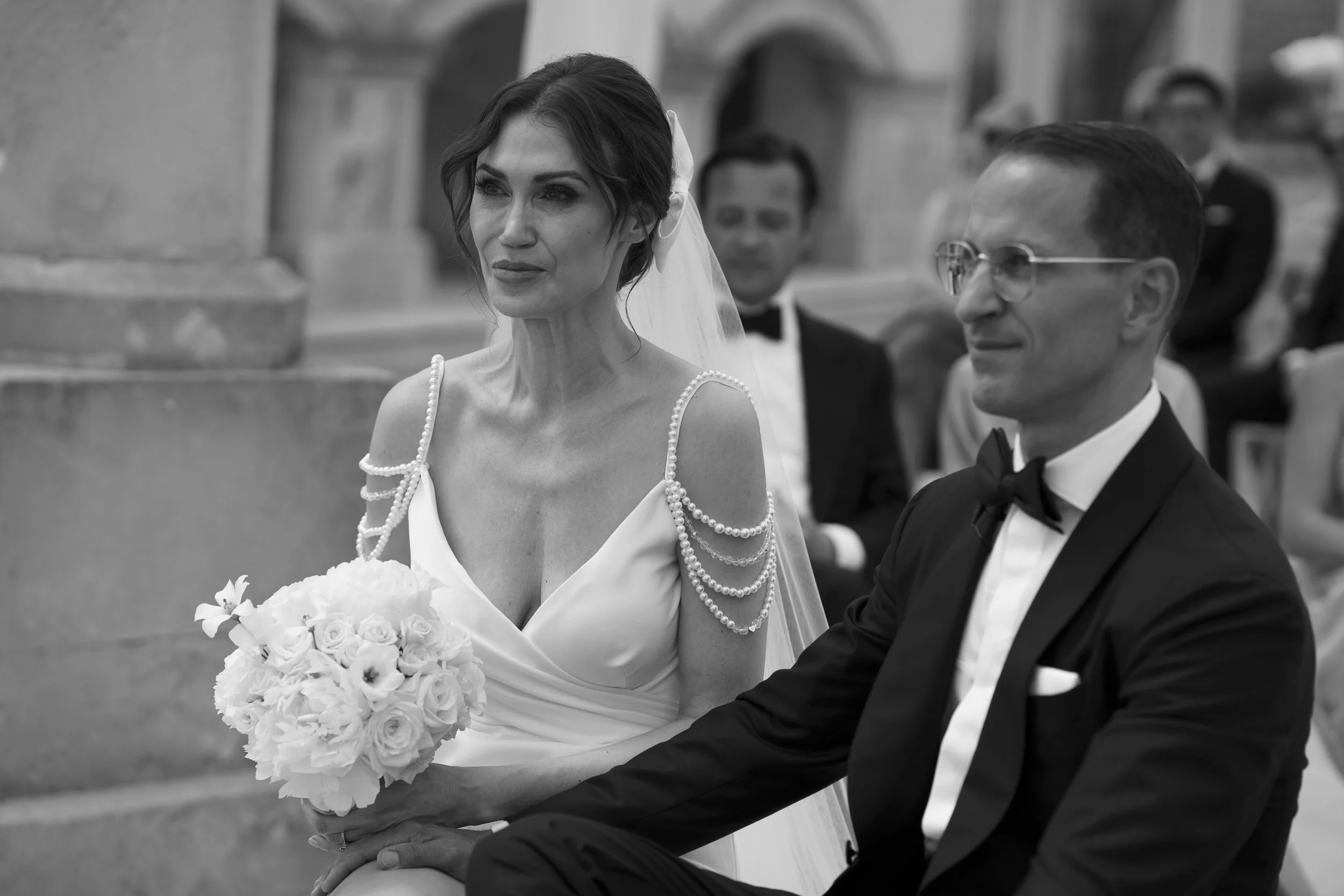 A black and white photo of a bride holding a bouquet of flowers, sitting next to a groom in a tuxedo, at a wedding ceremony.