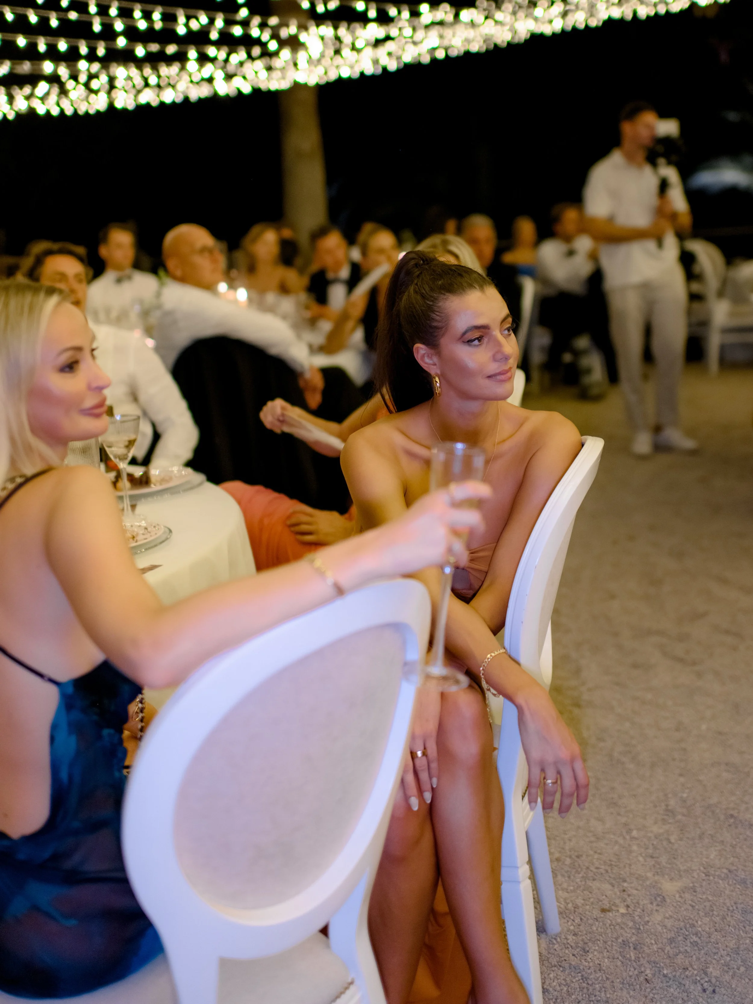 Guests seated at a formal event under string lights, with women in elegant dresses and a man standing with a camera in the background.