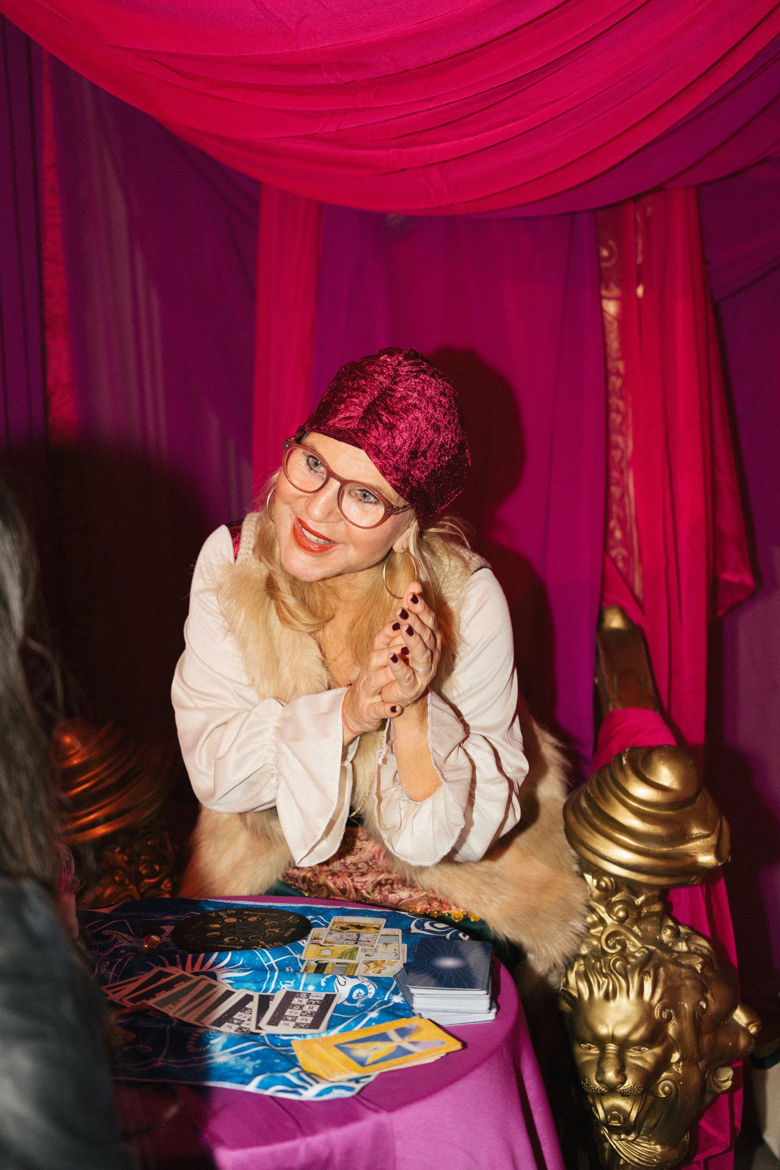 A woman with blonde hair, glasses, and red lipstick, dressed in vintage clothing, is smiling and sitting at a decorated table with tarot cards and a crystal ball, in a purple and pink draped setting.