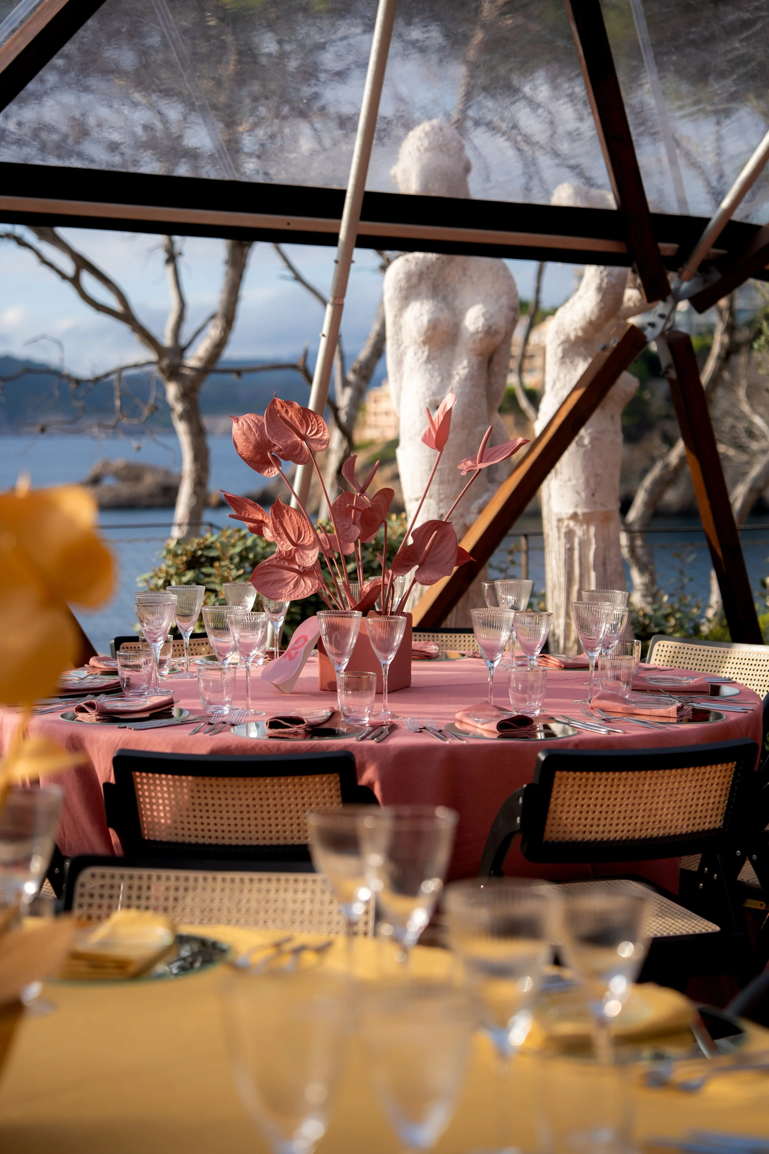 Elegant outdoor dining table set with pink tablecloth, pink floral centerpiece, glassware, and napkins, under a glass canopy with a scenic ocean view and trees in the background.