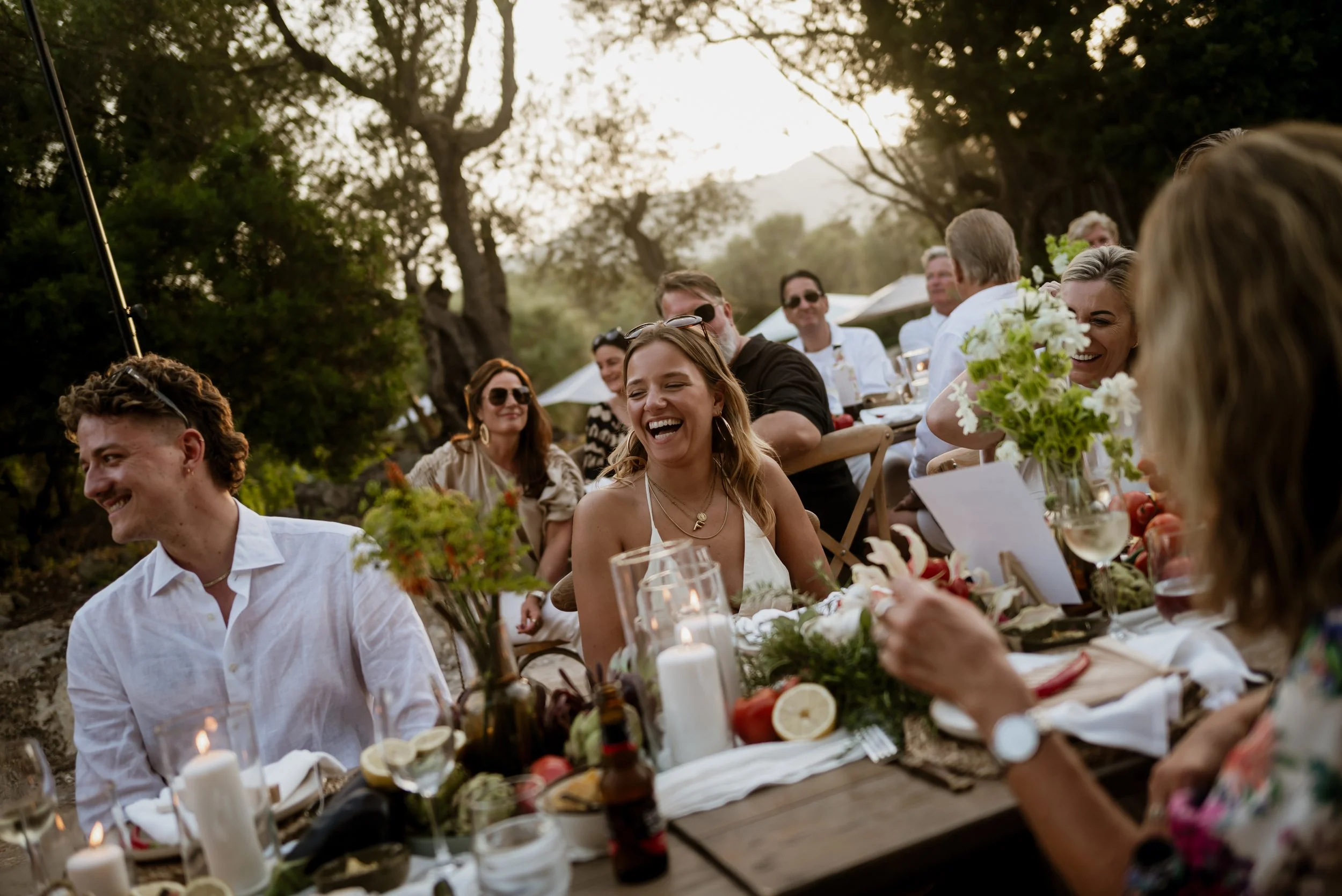 People sitting at a decorated outdoor dinner table, laughing and smiling, with trees and sunset in the background.