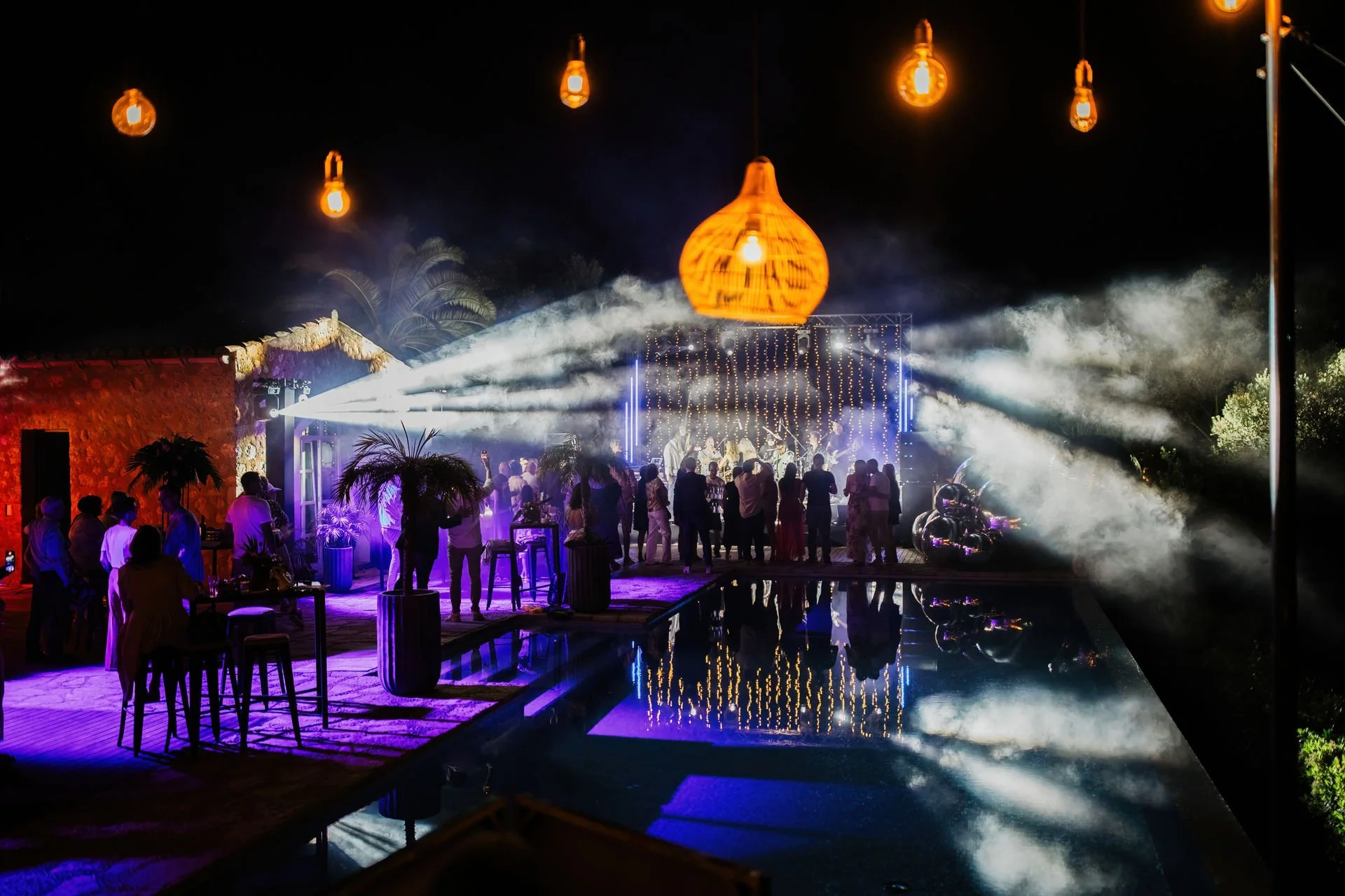 Nighttime outdoor party with people dancing and socializing under hanging lights near a pool, with a stage illuminated by spotlights and string lights.