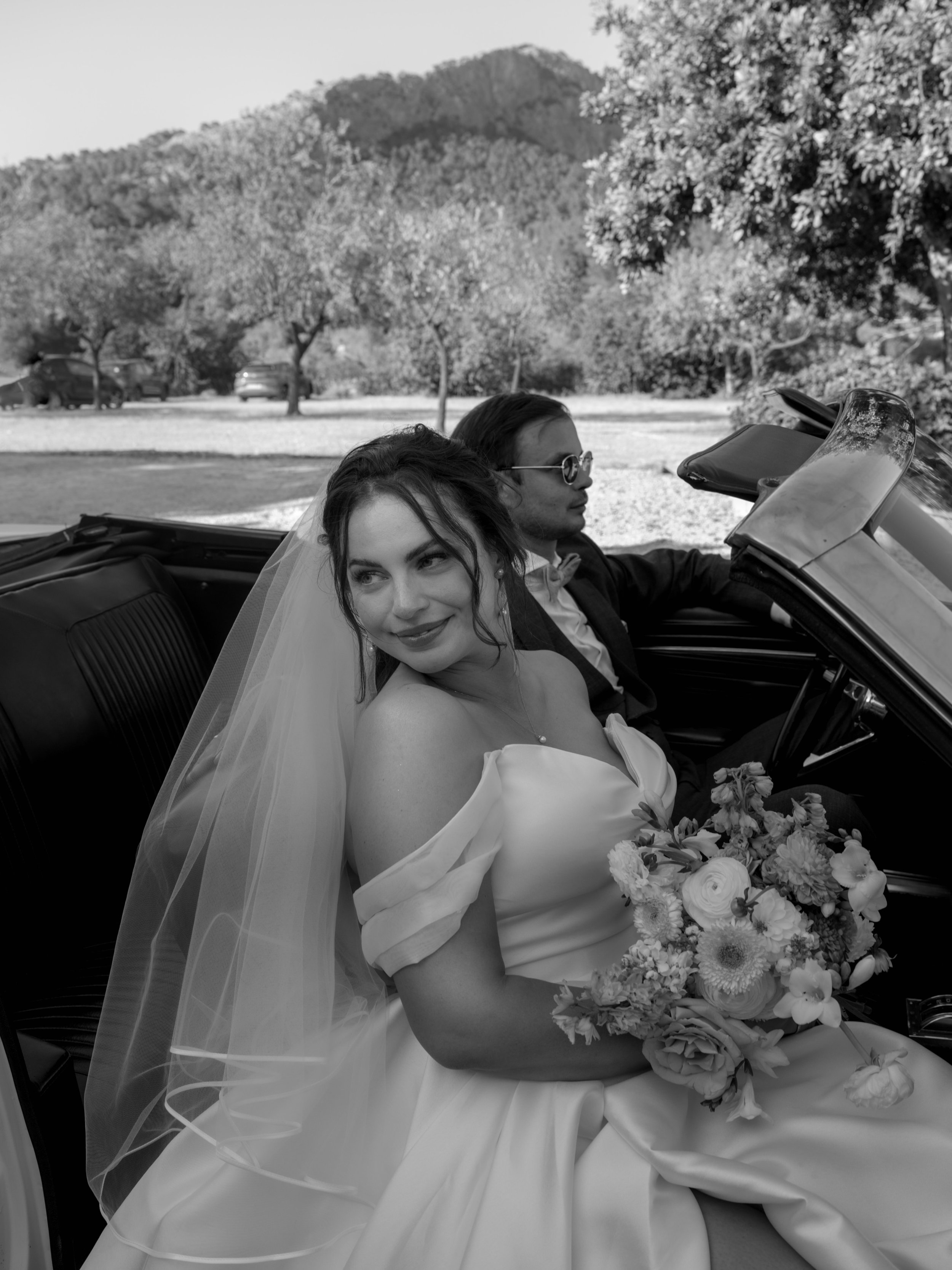 A bride in a wedding dress and veil sitting in a convertible car holding a bouquet, with a groom in a suit and sunglasses sitting beside her in a scenic outdoor location with trees and hills in the background.