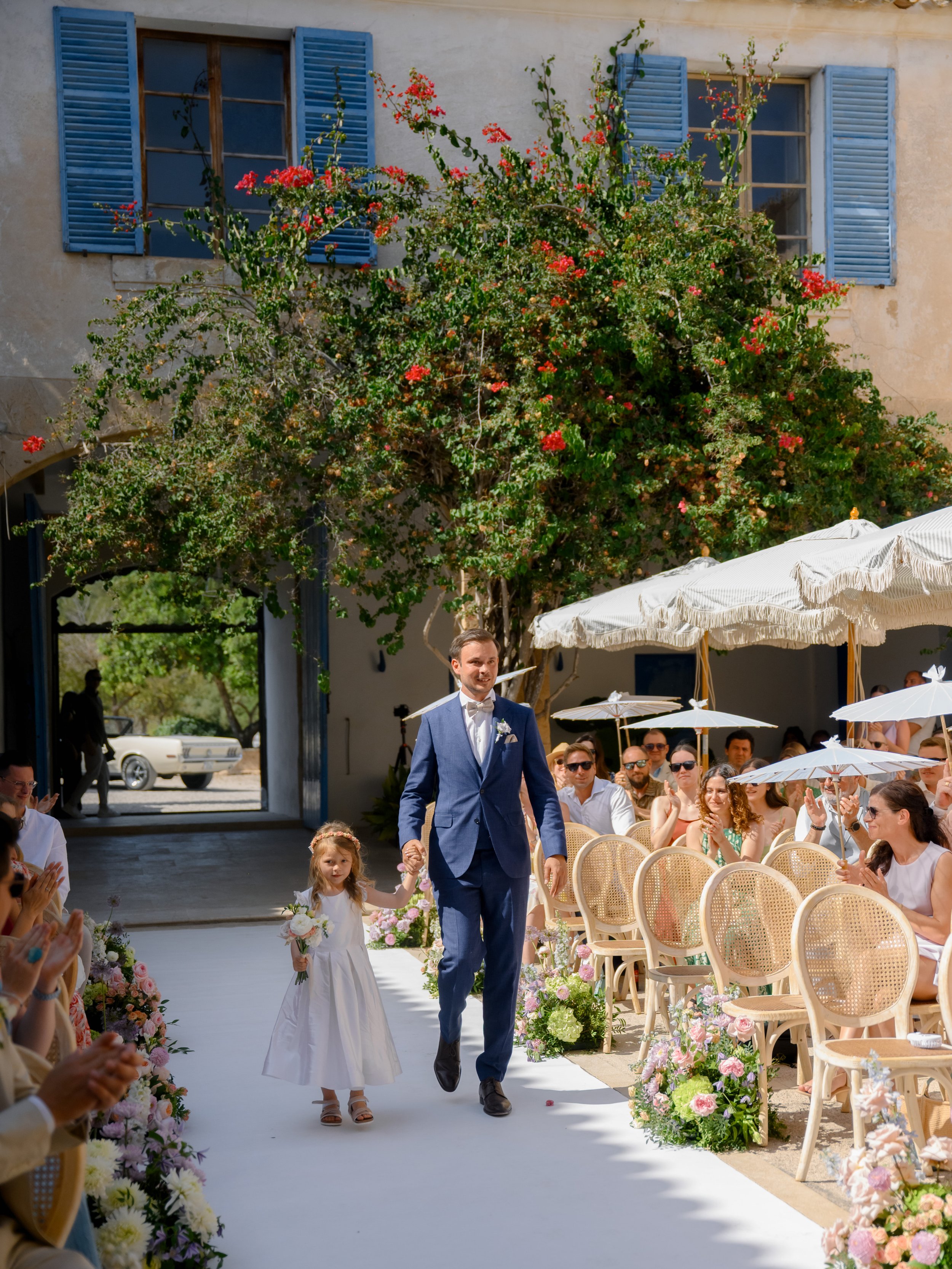 A wedding ceremony outdoors with a little girl and a man in a blue suit walking down the aisle. Guests seated on either side, some under white parasols, with a flowering tree and building with blue shutters in the background.