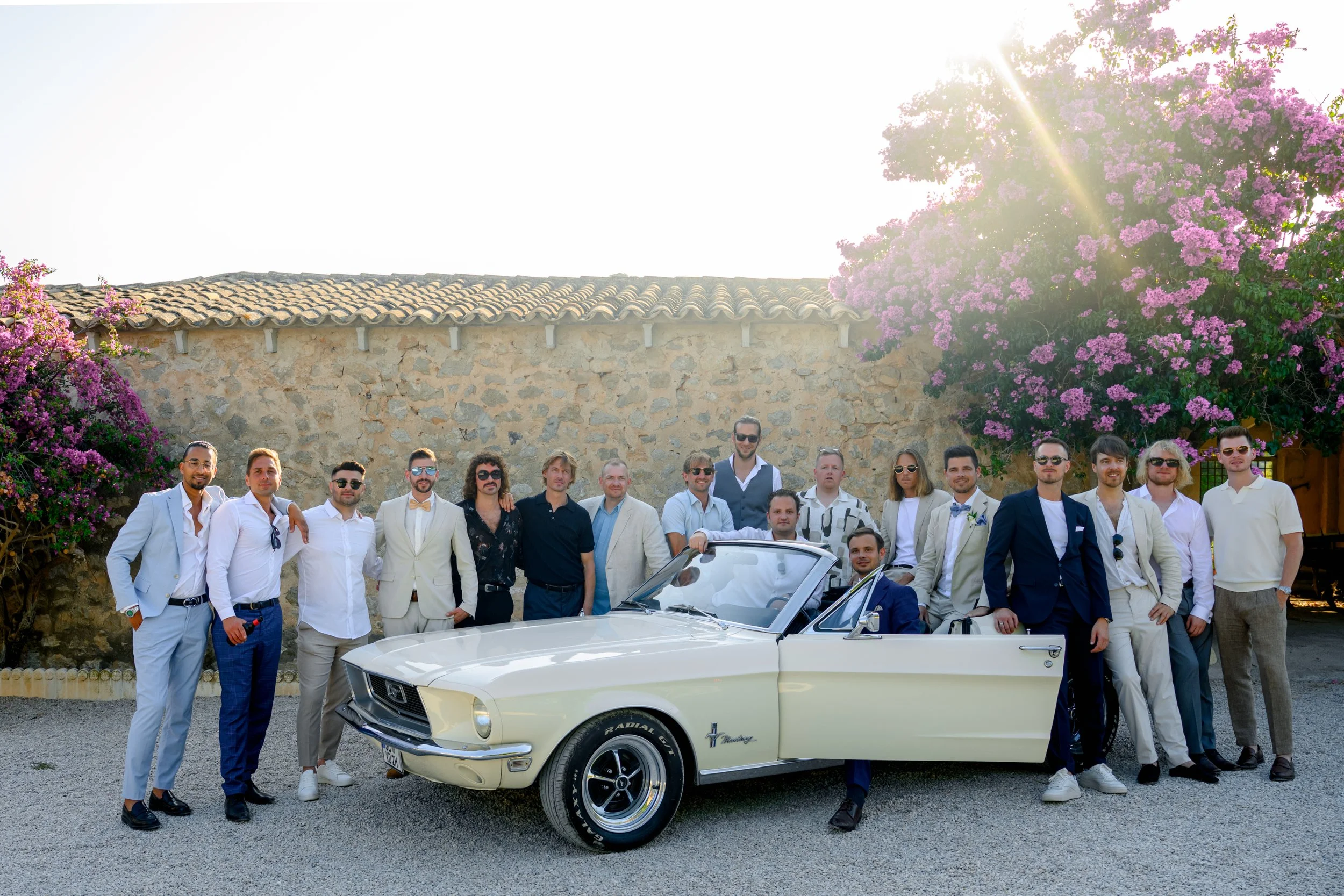 A group of people dressed in formal and semi-formal attire gathered around a vintage convertible car, with a stone building, pink flowering trees, and the sun shining brightly in the background.