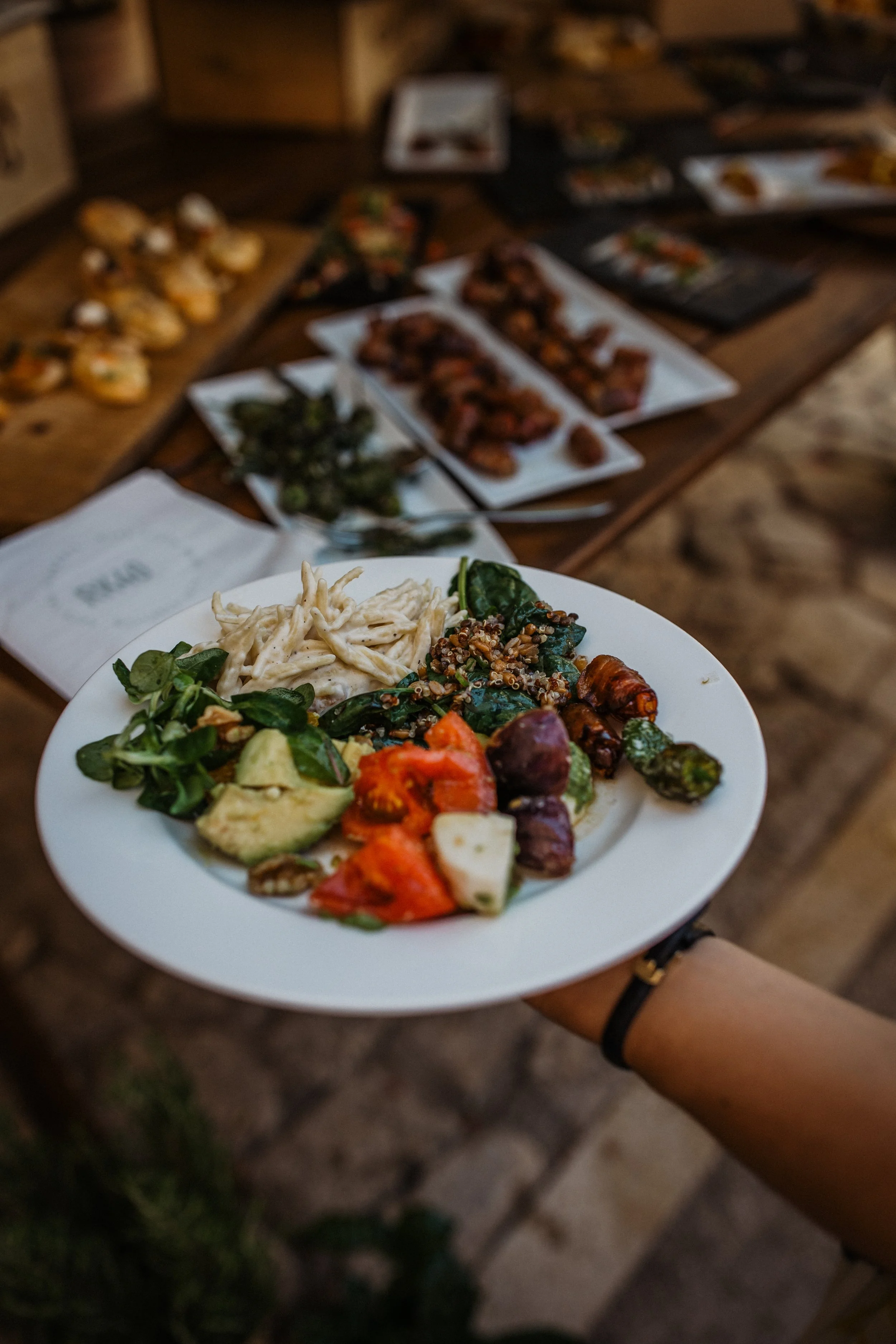 A white plate with assorted vegetables and pasta, held by a person wearing a black watch, with a wooden table in the background showcasing various appetizers and dishes.