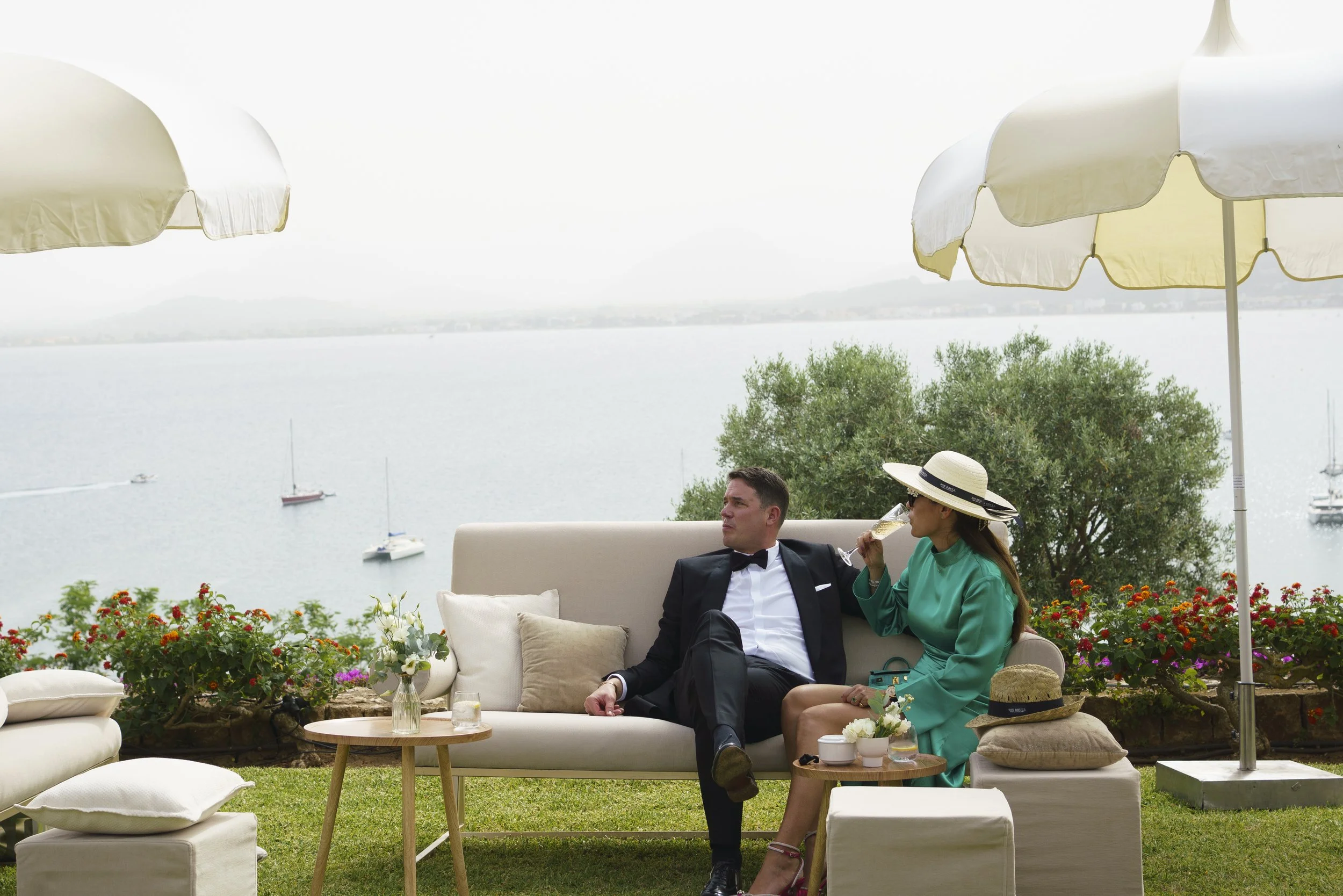 A couple sitting on a beige outdoor couch under umbrellas by a lake, with boats in the water and mountains in the distance. The man is dressed in a tuxedo and the woman in a teal dress and wide-brimmed hat, enjoying drinks.