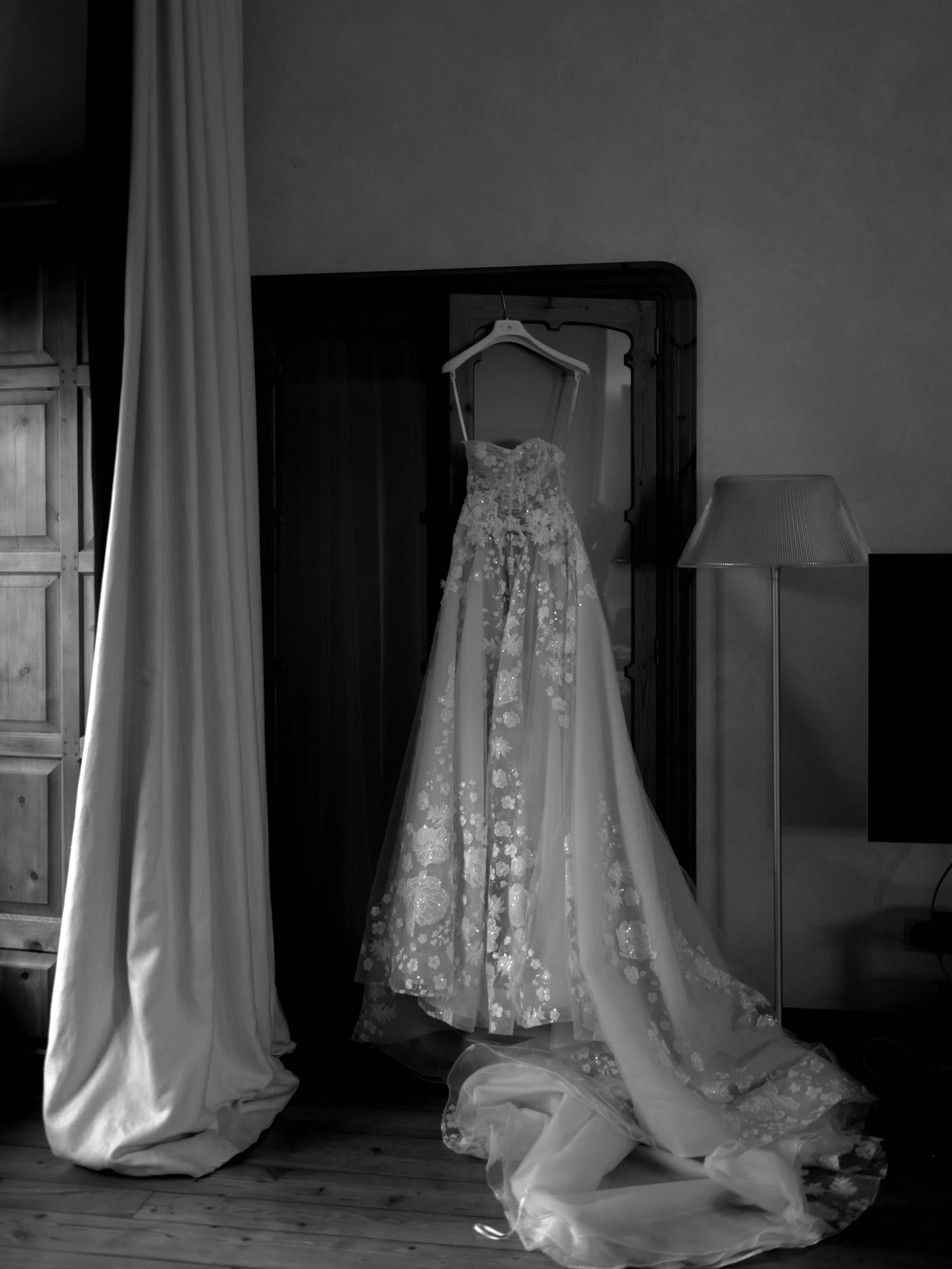 Wedding dress hanging on a hanger in a room, draped over a wooden armoire, with a floor lamp and curtains nearby.