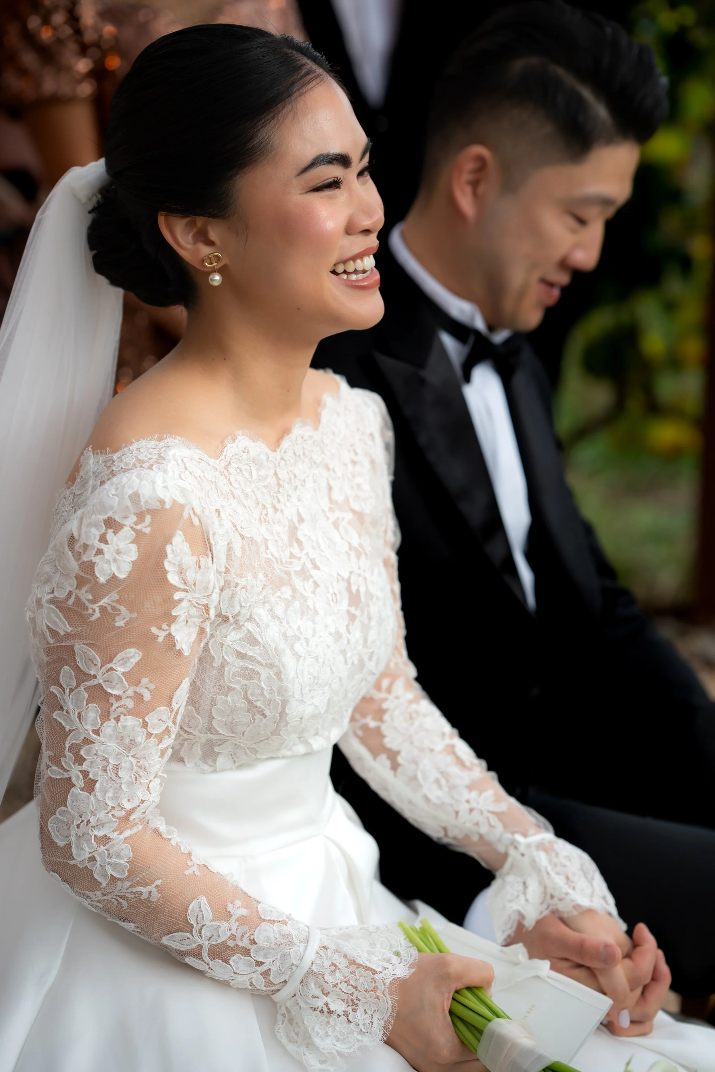 A bride in a white lace wedding dress and pearl earrings smiling, seated next to a groom in a black tuxedo, holding hands, during a wedding ceremony.