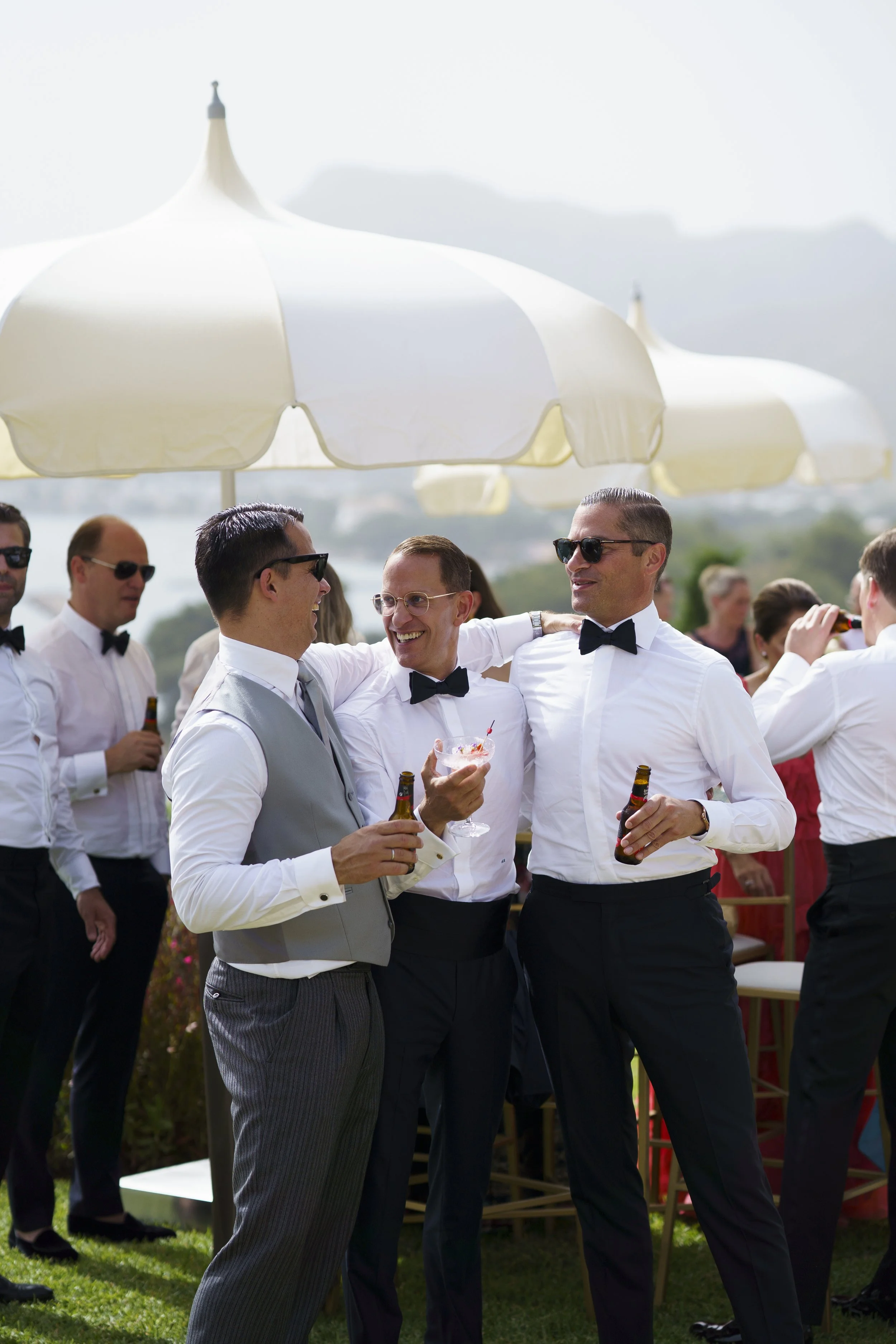 Group of men in formal attire enjoying drinks and socializing outdoors at a celebration, possibly a wedding or formal event, under large umbrellas with scenic mountains in background.