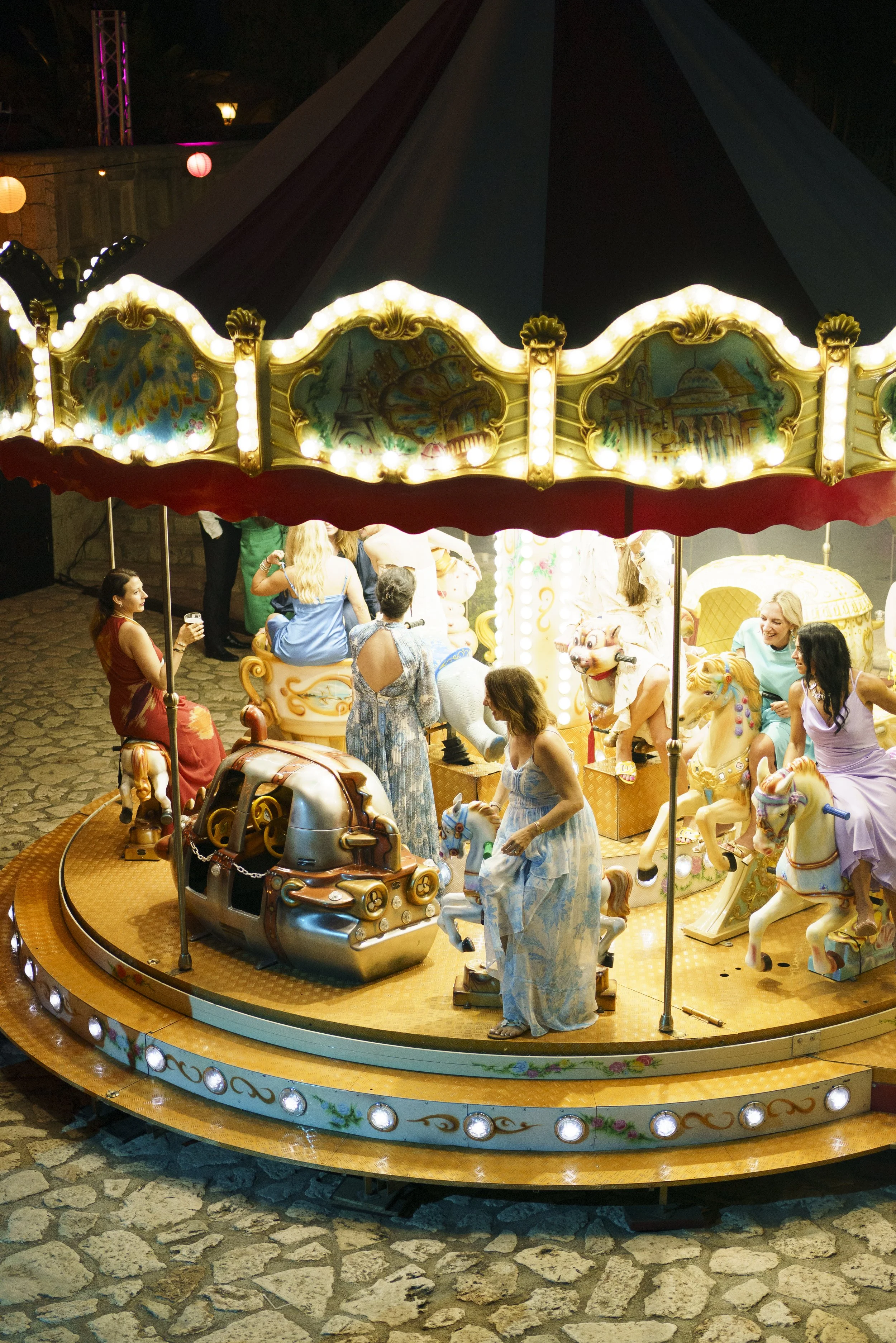 Colorful carousel with children and adults riding and standing near, decorated with bright lights and vintage-style artwork, set outdoors at night on cobblestone ground.