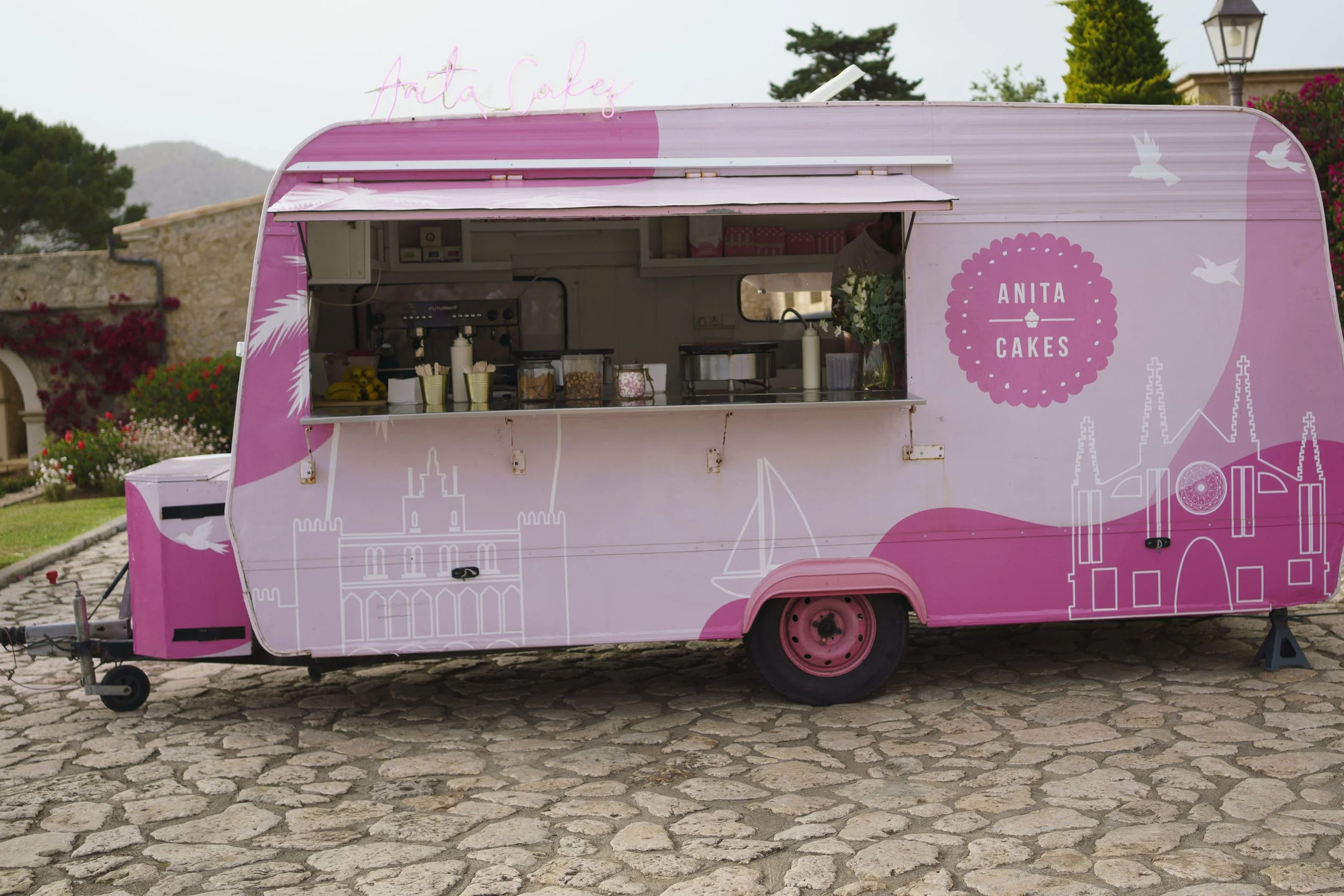 Pink food truck with the words "Anita Cakes" and illustrations of a castle and birds, parked on cobblestone ground with a garden and trees in the background.