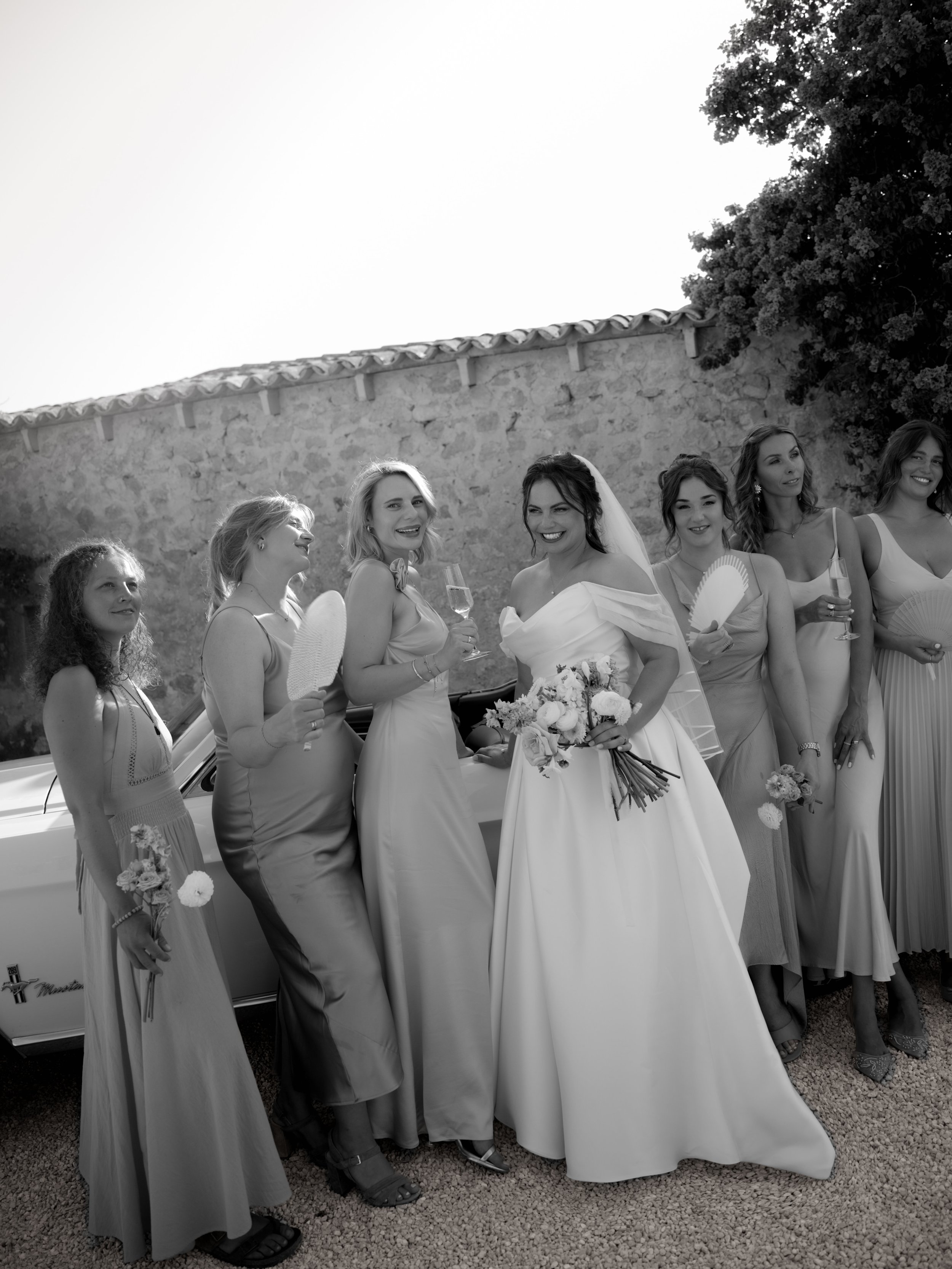 Black and white photo of a bride with six bridesmaids outdoors, smiling and holding flowers and fans.