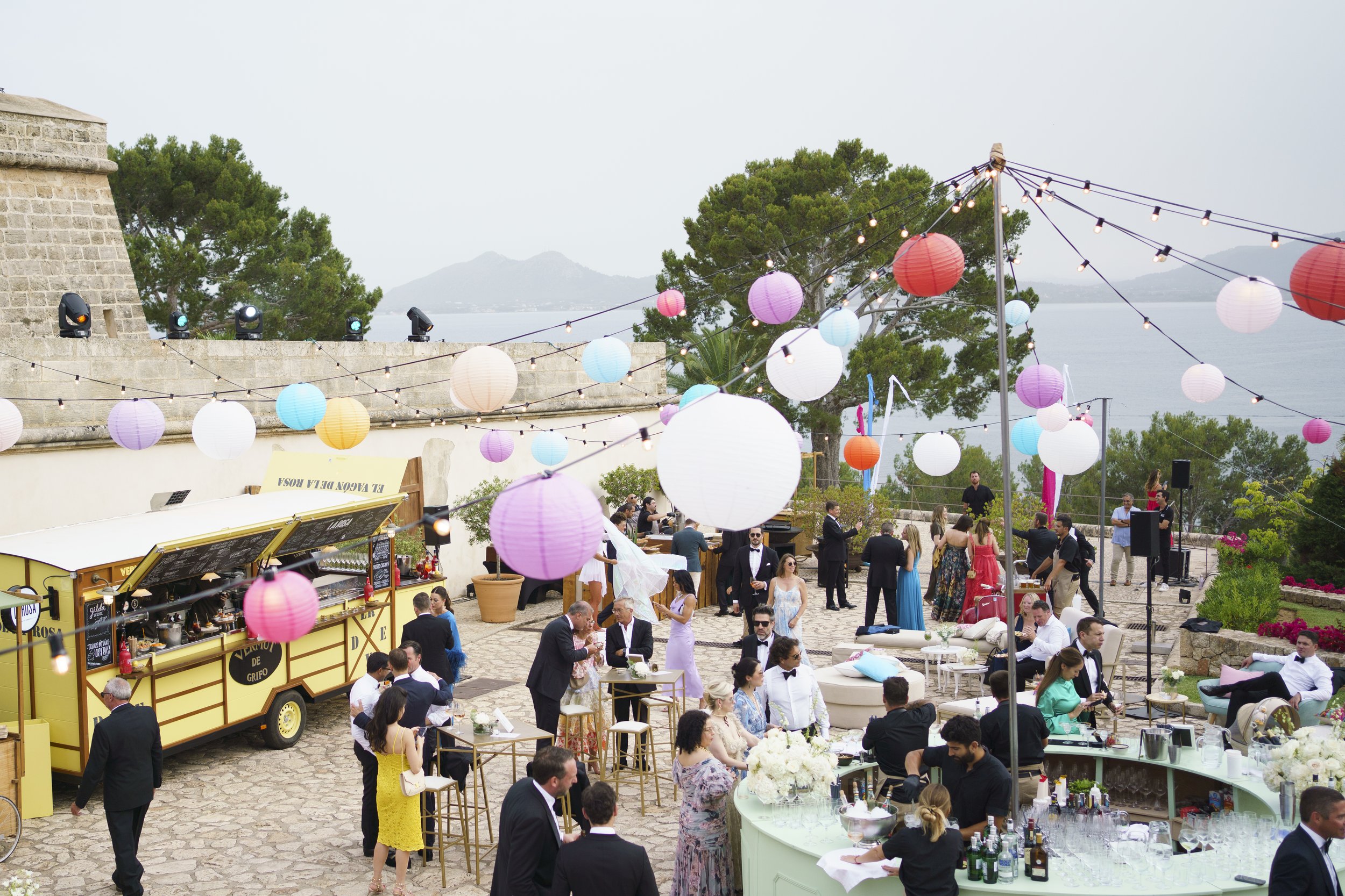 Outdoor wedding reception with guests, string lights, paper lanterns, a bar, and a scenic view of trees and water in the background.