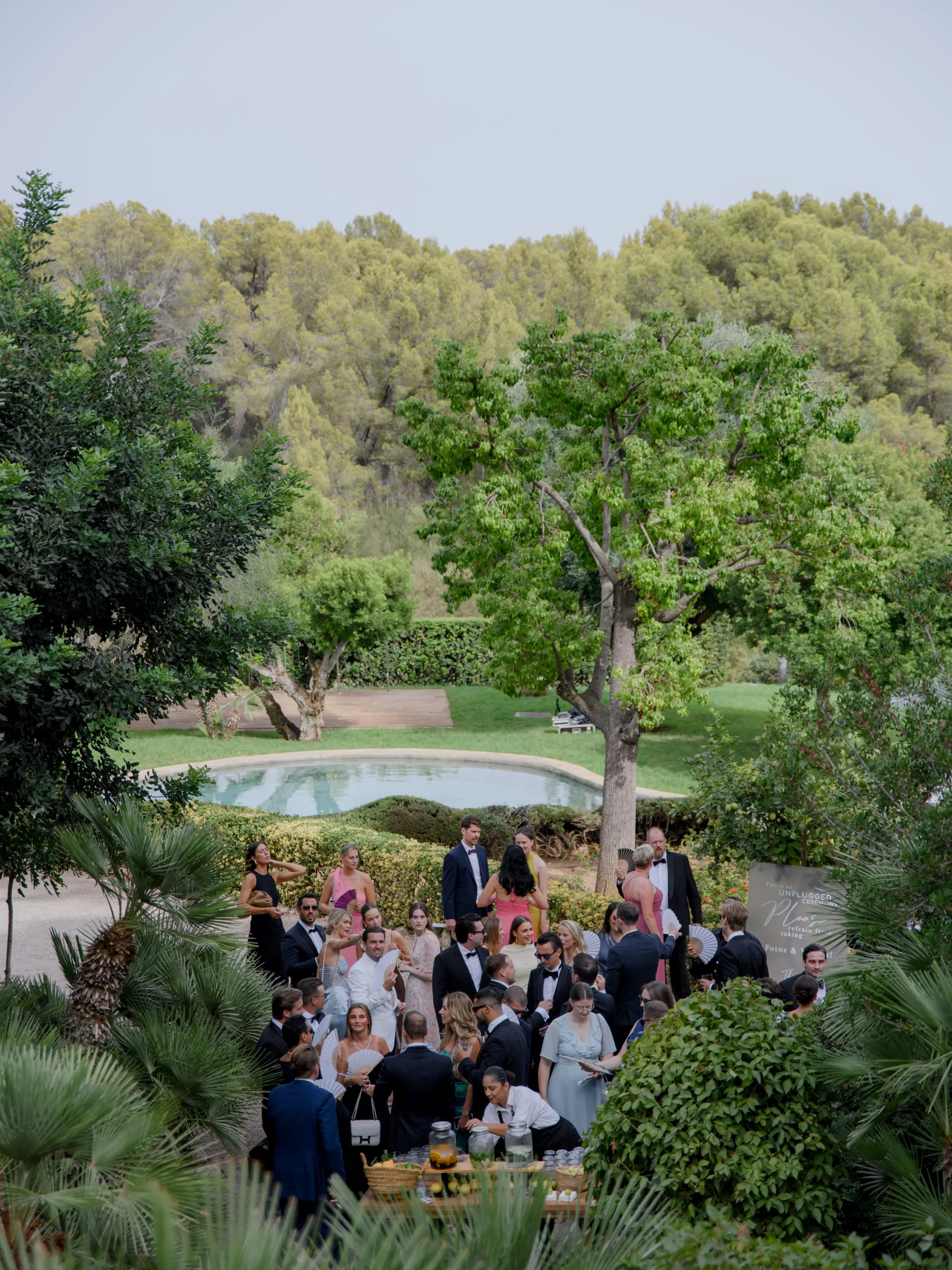 A large outdoor gathering of people dressed in formal attire, with some holding fans, set in a green park with trees, a small pond, and lush foliage.