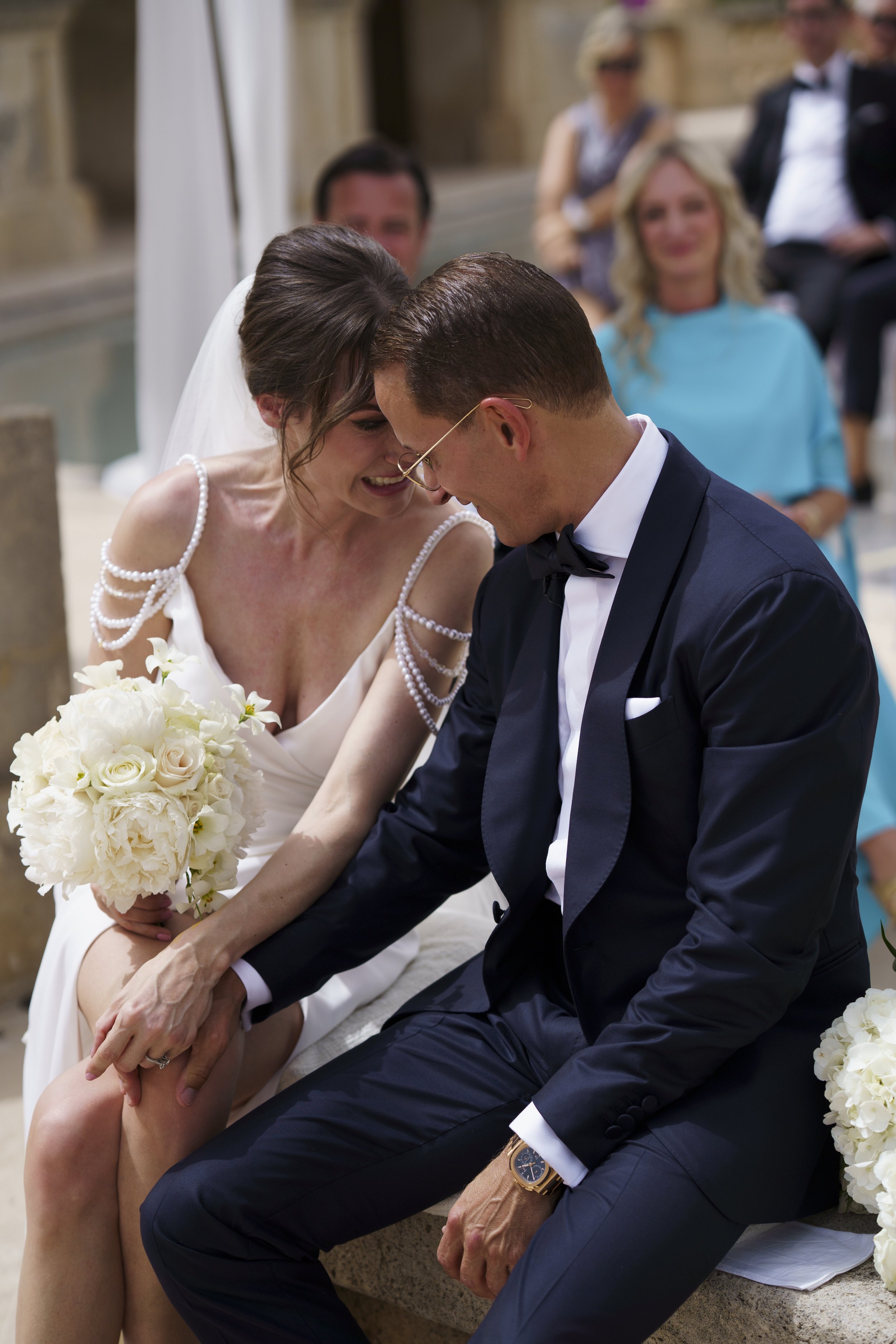 A bride and groom sitting close together, smiling, during their wedding ceremony, with guests watching in the background.