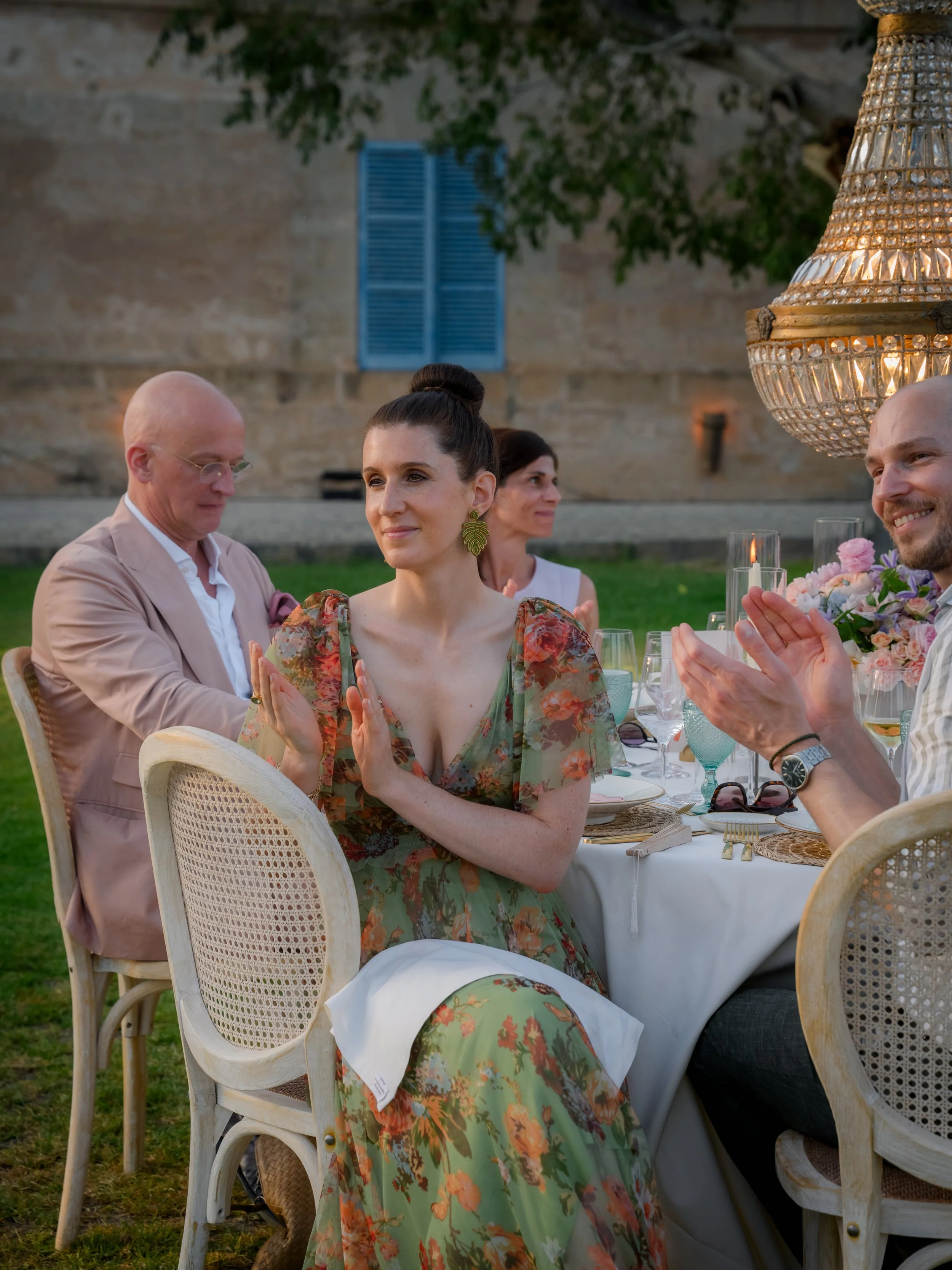 A woman in a floral dress with green and orange hues is sitting at an outdoor dinner table, clapping, with a man in a striped shirt also clapping beside her. Other guests are seated around the table, with a building and greenery in the background, an