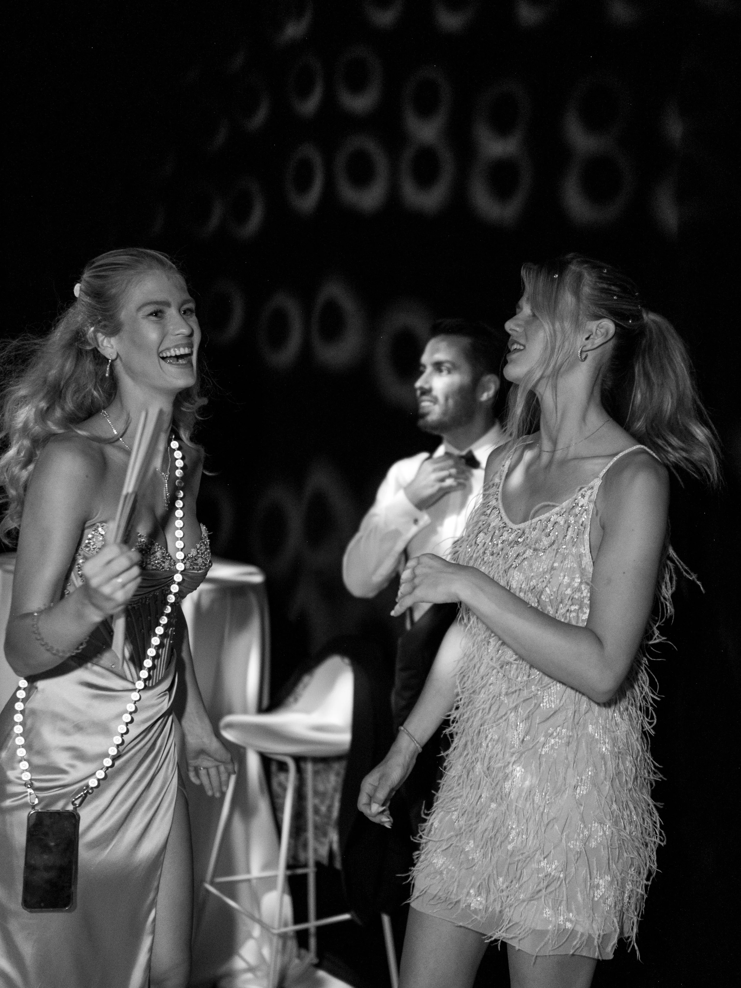 Three people at a party: two women dressed in glamorous evening wear are conversing, one smiling and holding chopsticks and a necklace, and the other looking at her with a smile. A man in the background, dressed in a white shirt with a bow tie, appea