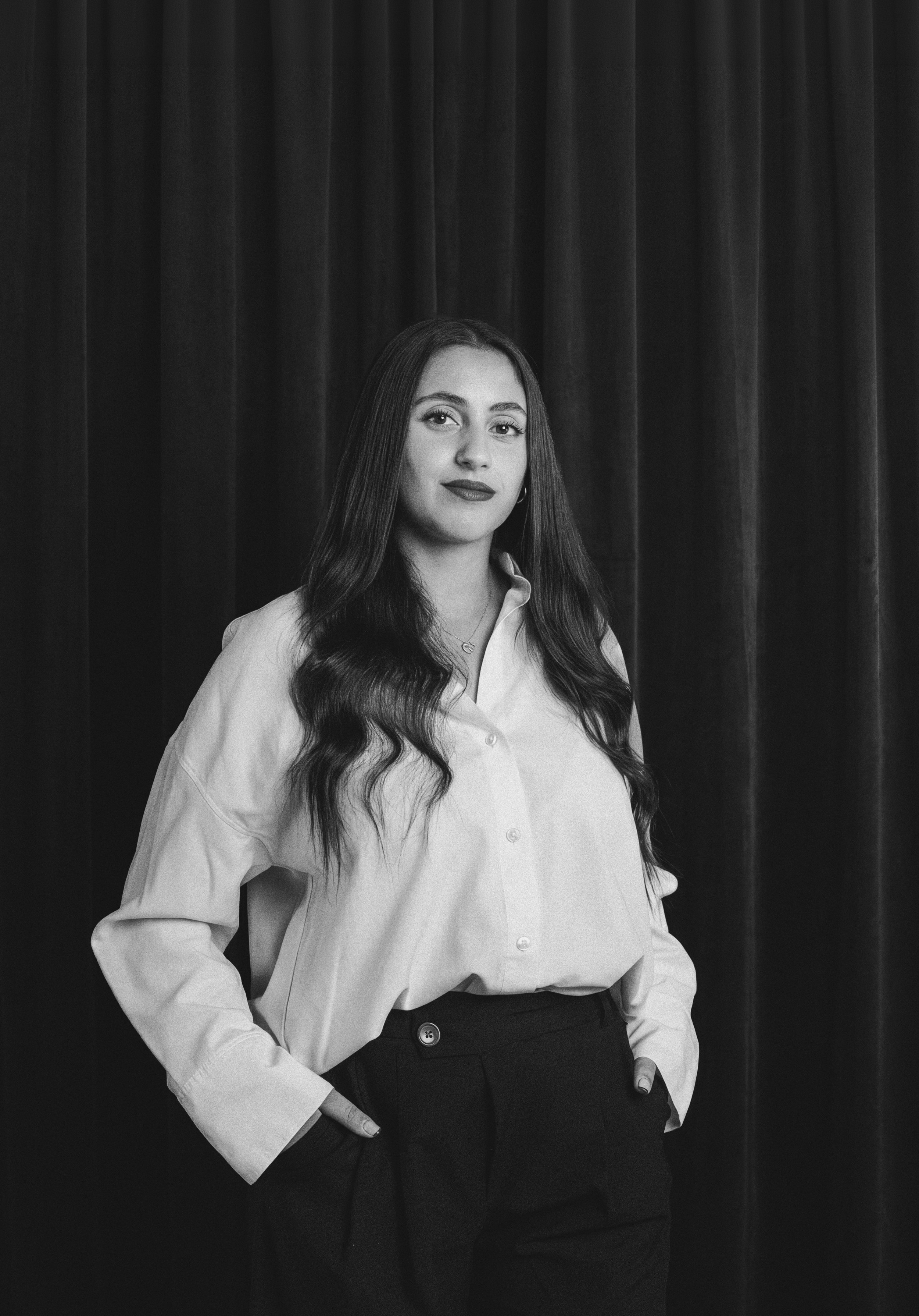 A portrait of a woman with long wavy hair, wearing a light-colored shirt and dark trousers, standing in front of a dark curtain.