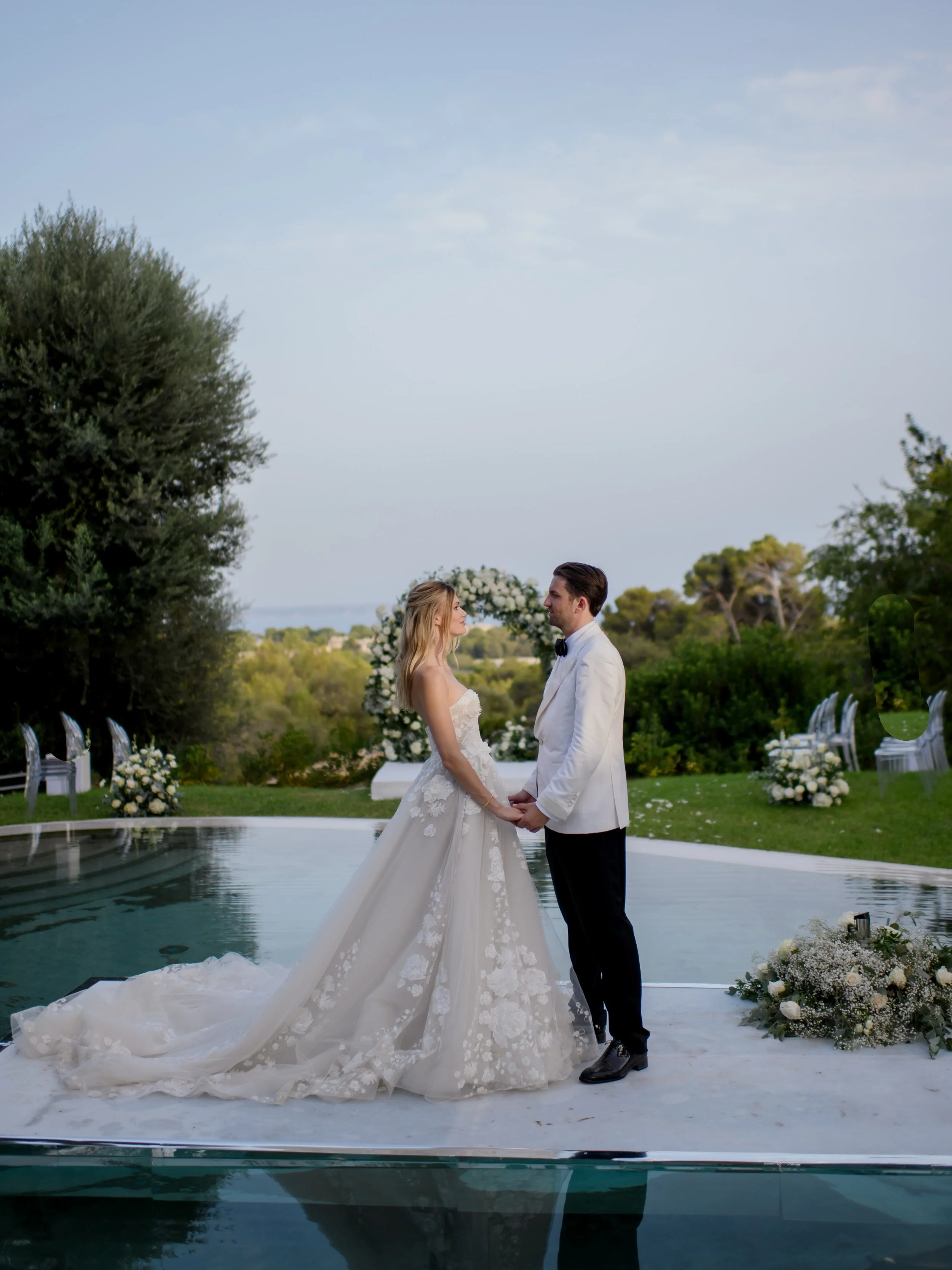 Bride and groom exchanging vows during wedding ceremony outdoors, standing in front of a pool with decorated surroundings and greenery.